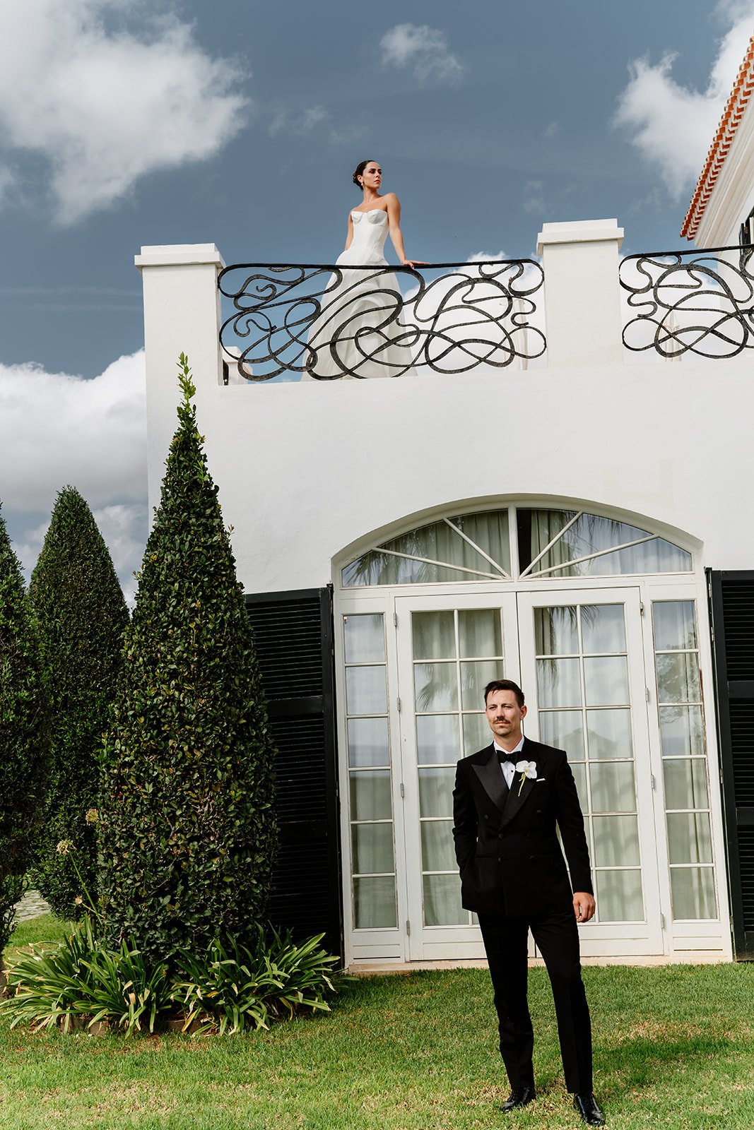 Bride on balcony and groom standing below villa during destination wedding in Menorca