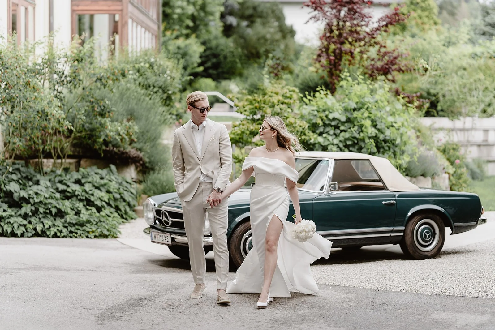 Braut und Bräutigam spazieren Hand in Hand an einer Mercedes Pagode vor dem Hotel Lengbachhof vorbei.