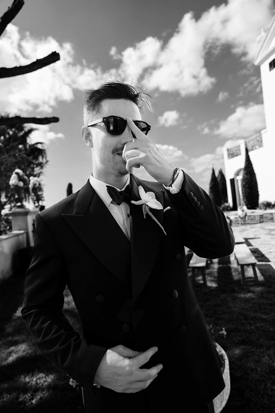 Black and white portrait of a groom adjusting his sunglasses in a modern tuxedo at a Mediterranean villa wedding
