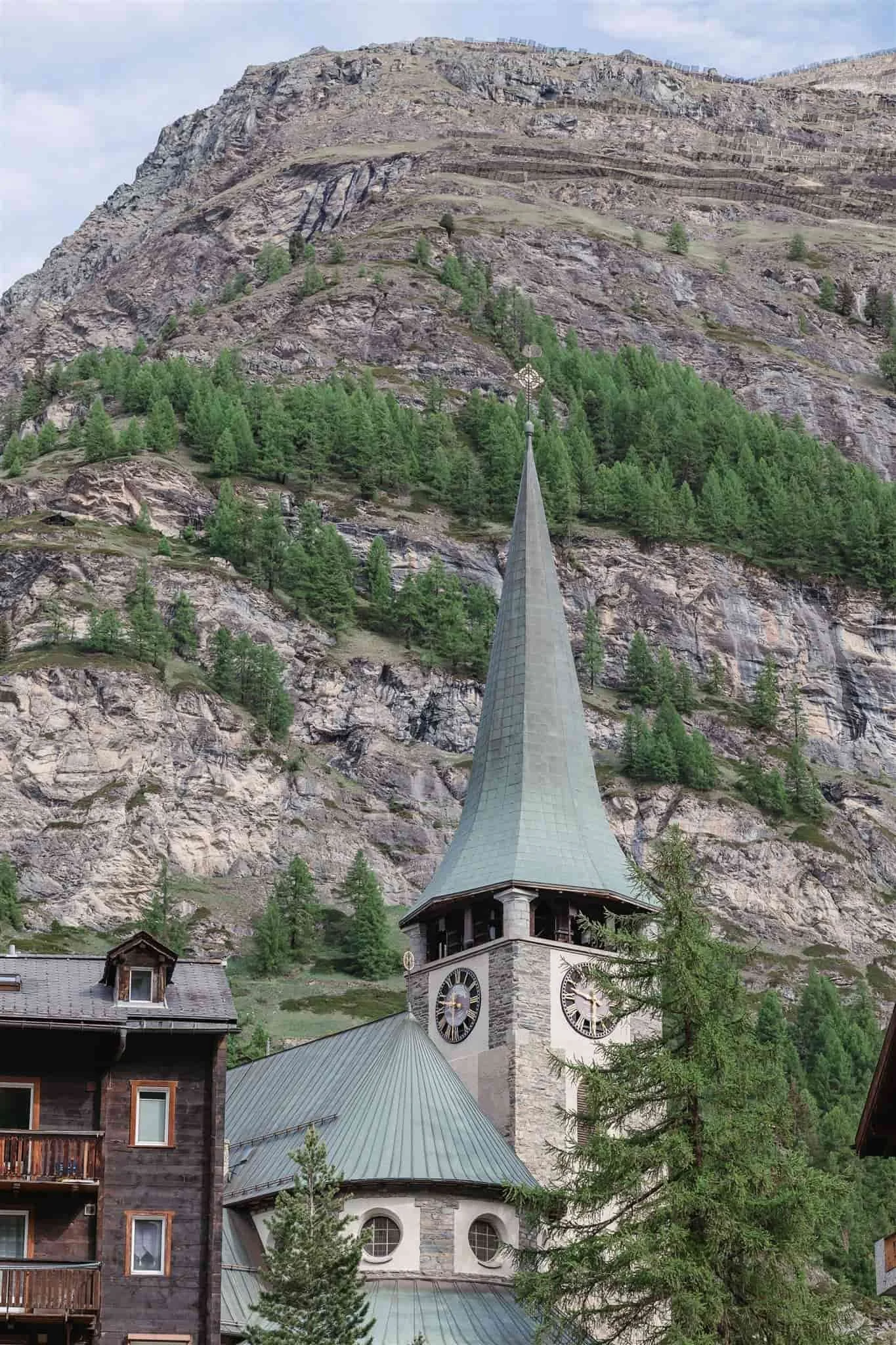 A church with a tall steeple and clock faces stands before a forested mountain slope, captured by a Portugal wedding photographer.