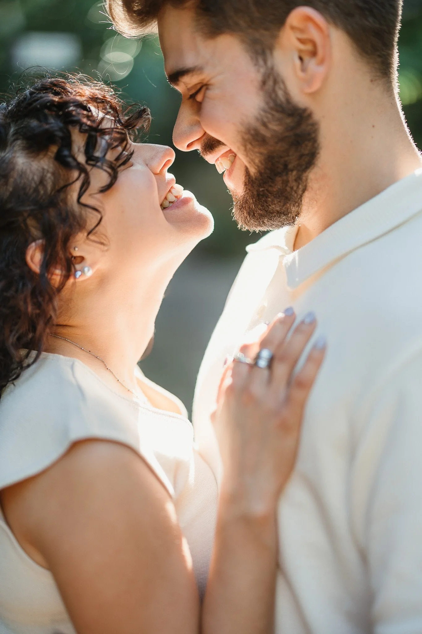Engagement Session: A man and woman face each other closely, smiling and touching foreheads, in an outdoor setting with sunlight filtering through trees.