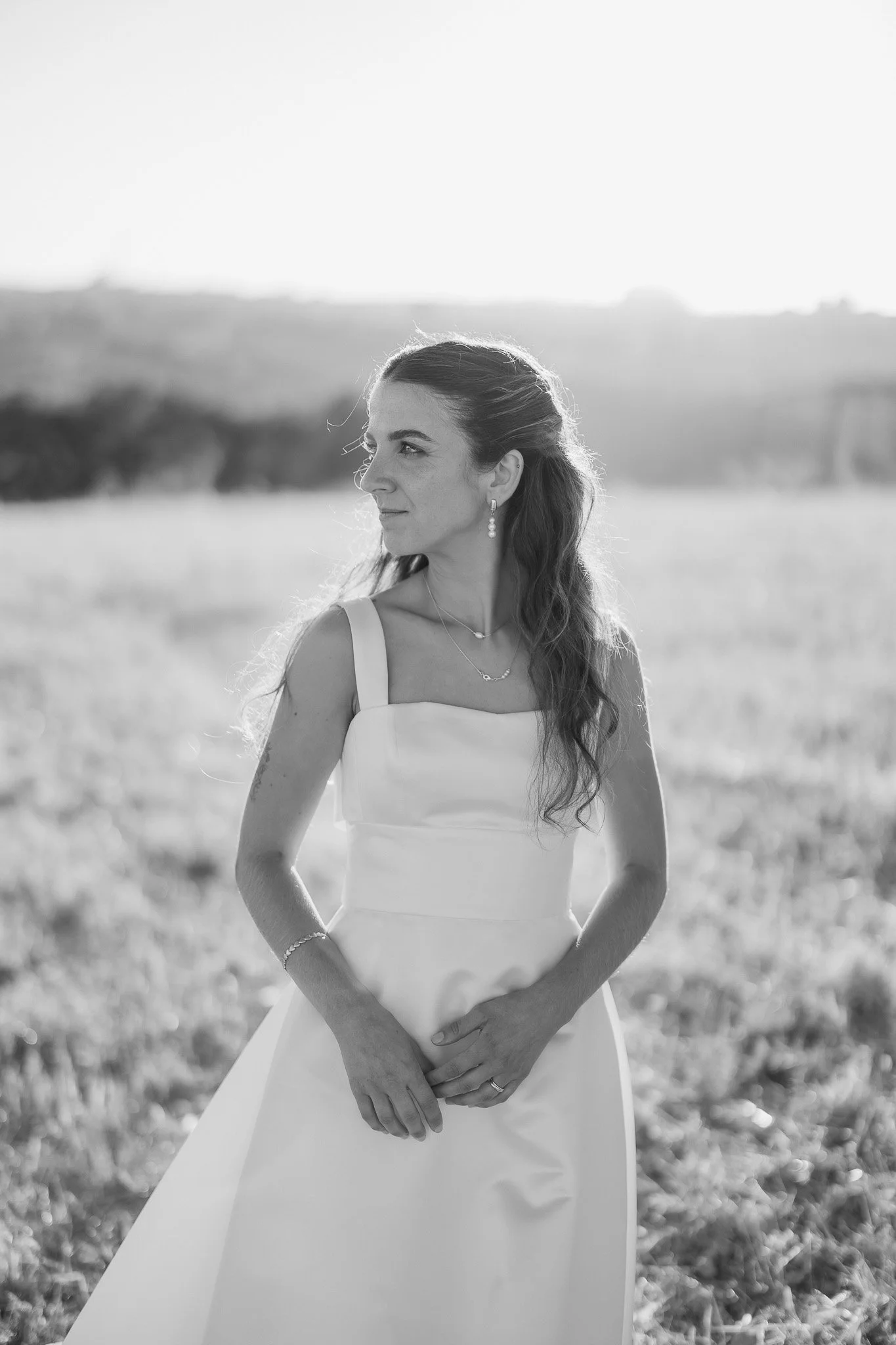 Noiva com vestido branco em pé num campo ao ar livre, olhando para o lado com expressão serena, em preto e branco. Fotógrafo de Casamento em Portugal.