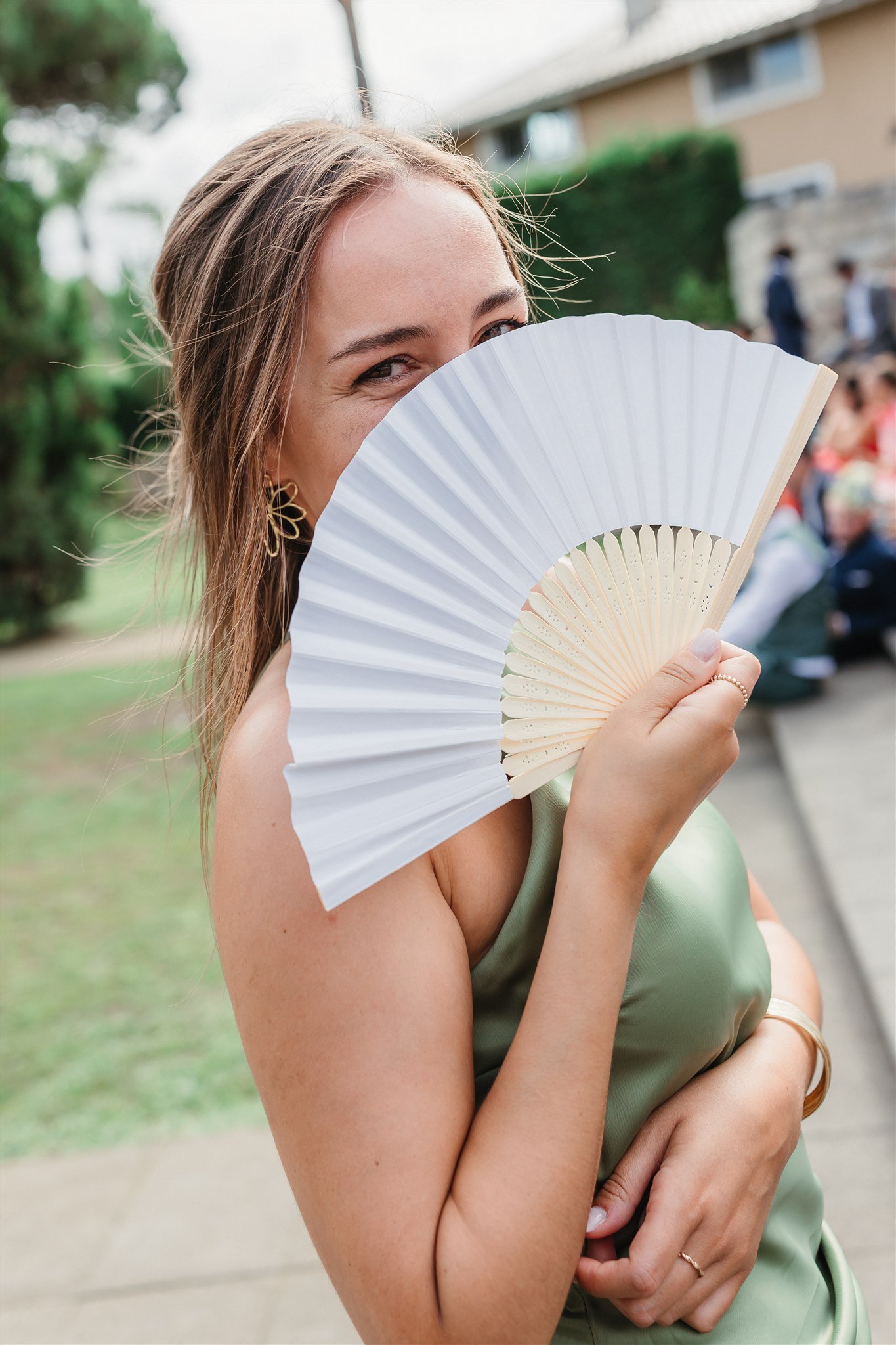 Emotional wedding moment in Portugal: A woman holding a white folding fan partially covering her face, smiling and wearing a green satin dress, outdoors with blurred background of people and greenery.