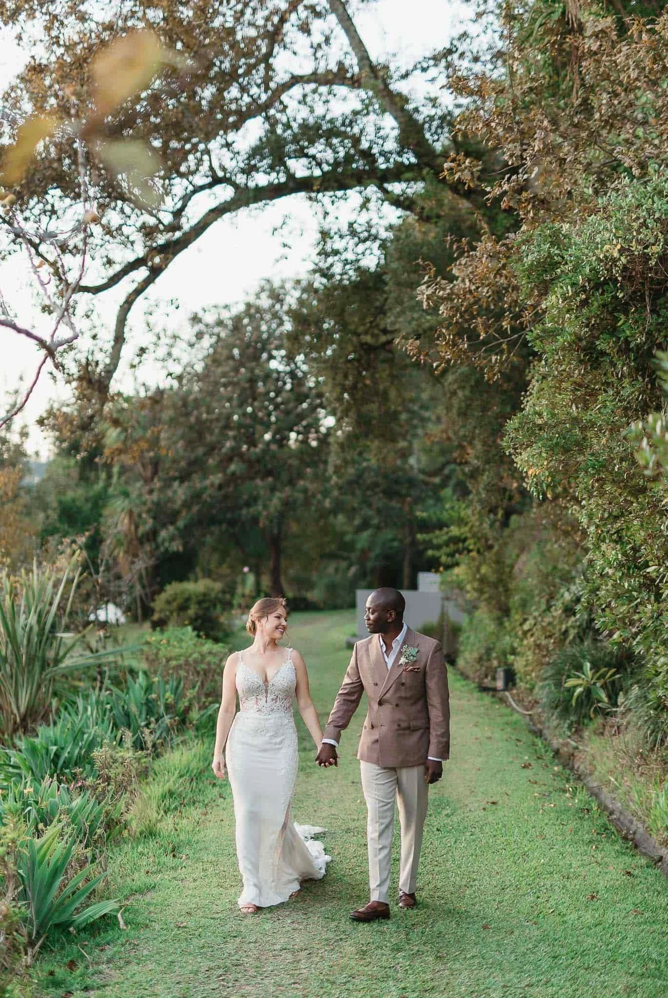 A newlywed couple walking hand-in-hand on a lush green garden path with trees and plants around, smiling at each other.
