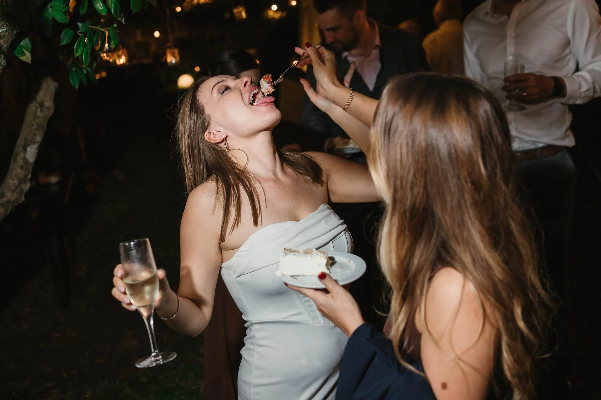 Two women at a party; one feeds cake to the other, who laughs with champagne—captured by a Portugal Wedding Photographer.