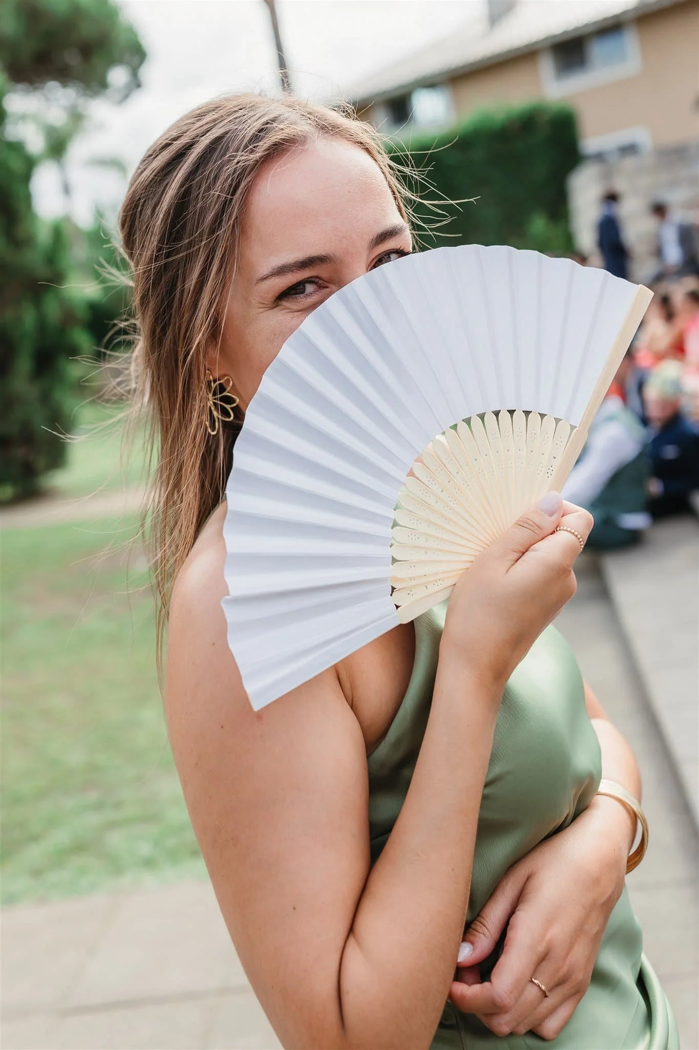 Young woman on a porch holding a white folding fan partially covering her face, smiling with eyes visible, wearing earrings and a green dress.