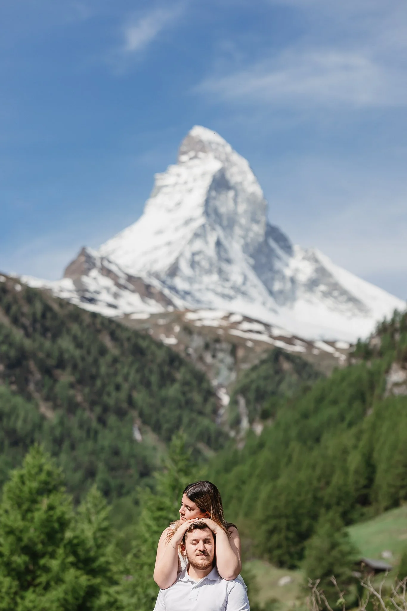 A couple stands outdoors with a snow-capped mountain in the background in Zermatt Switzerland, the woman resting her head on the man's shoulder.