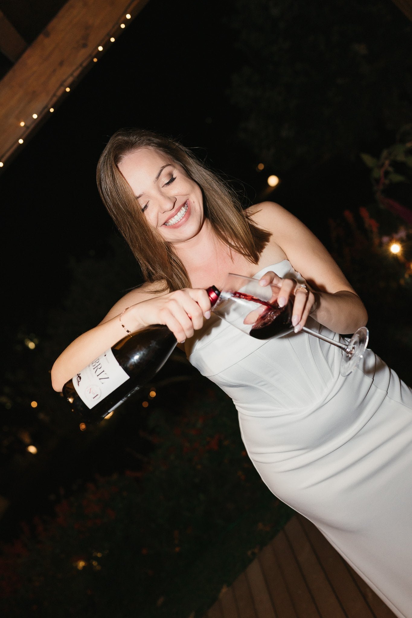 A woman wearing a white strapless dress, smiling with eyes closed, pouring red wine into a wine glass at an outdoor nighttime event.