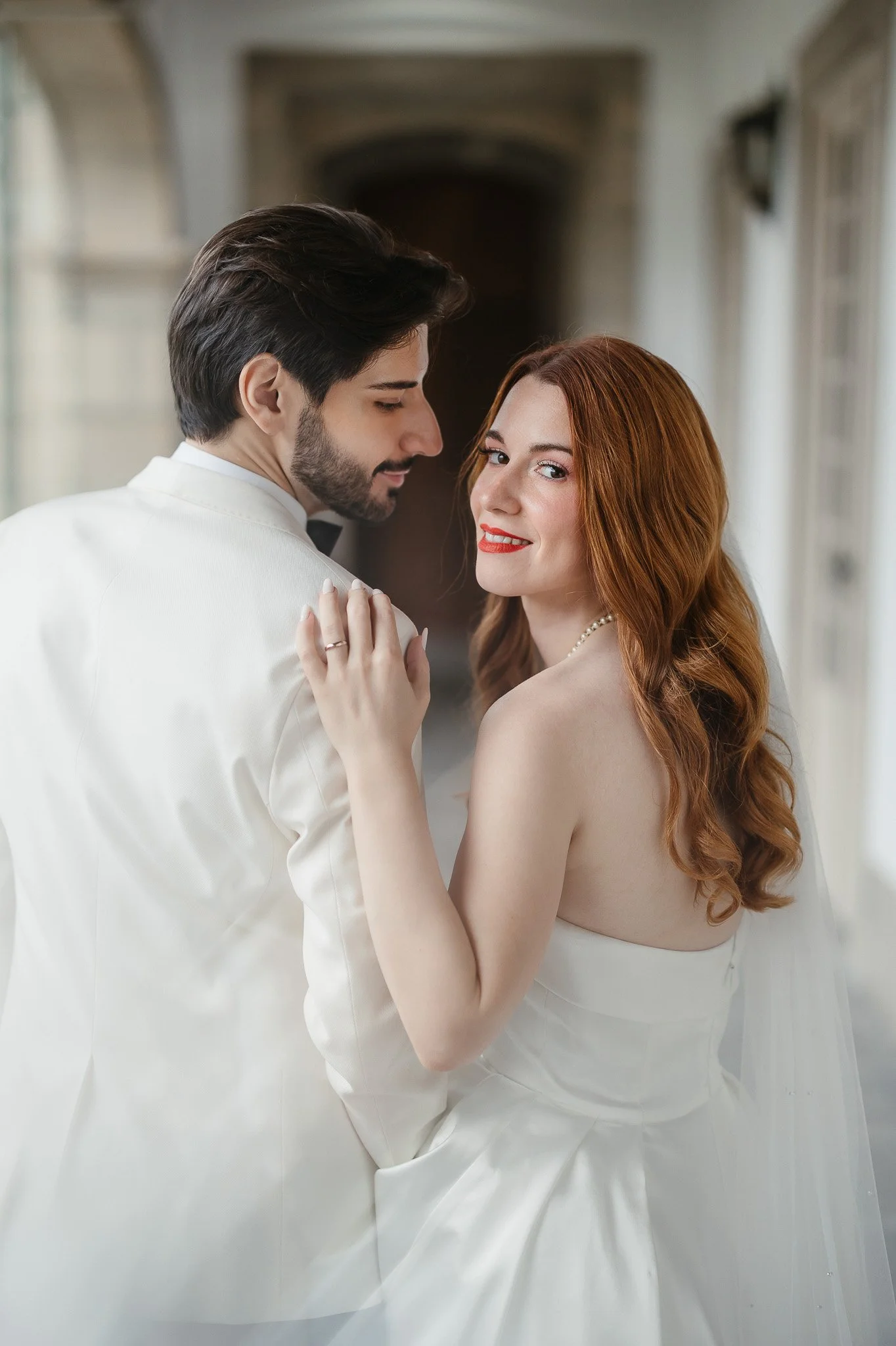 Emotional wedding moment in Portugal: A bride and groom on their wedding day, close-up portrait indoors, the bride with red hair and the groom with dark hair and beard, both in white wedding attire. Editorial Style Wedding