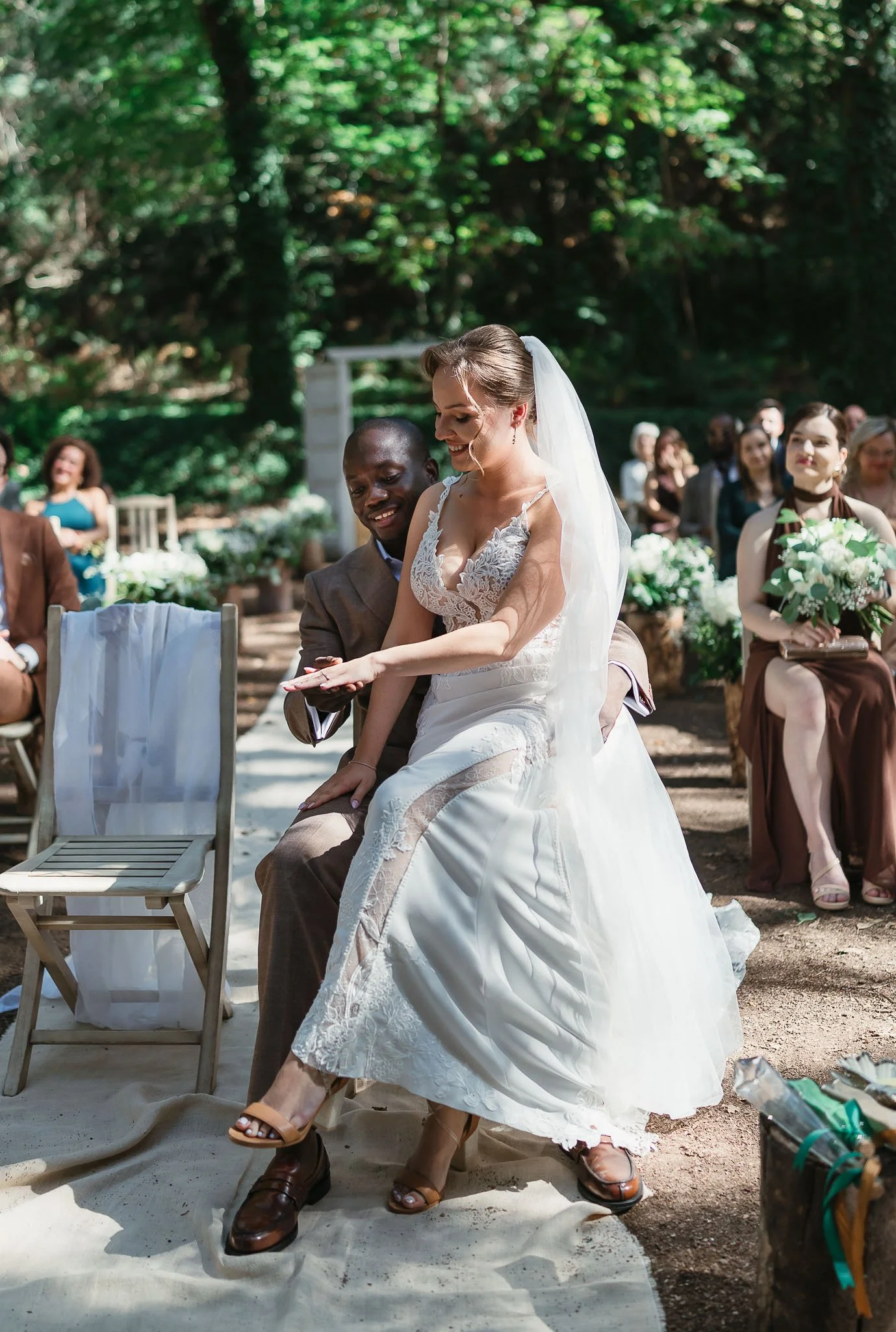 A bride in a white lace wedding dress and veil sitting on a man's lap during a wedding ceremony outdoors. Guests seated behind them in a forest setting, some holding bouquets, watching and smiling.