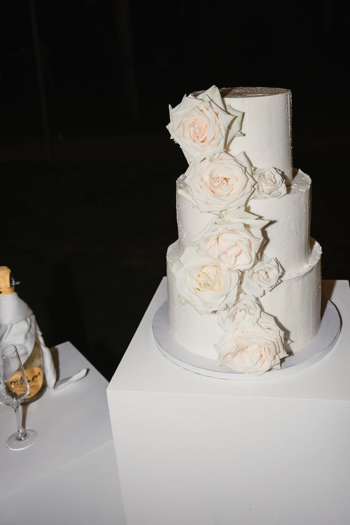 A three-tier white wedding cake with roses, captured by Portugal Wedding Photography beside champagne and a glass.