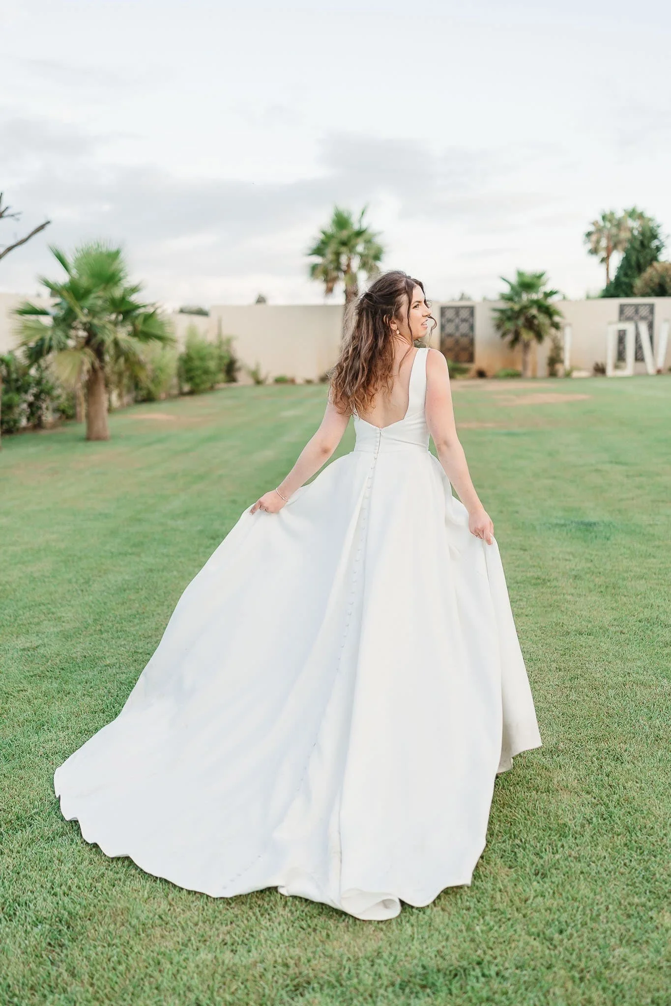 A woman in a white wedding dress standing on a lush green lawn with palm trees in the background. Quinta do Éden, Aveiro, Portugal