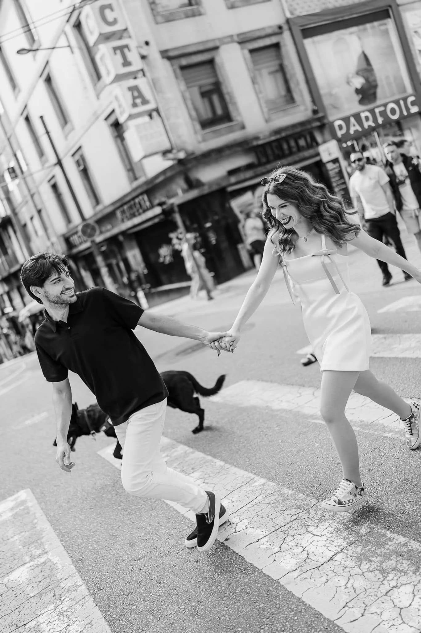 A smiling couple holds hands and runs across a city crosswalk with their dog, captured by a Portugal wedding photographer.