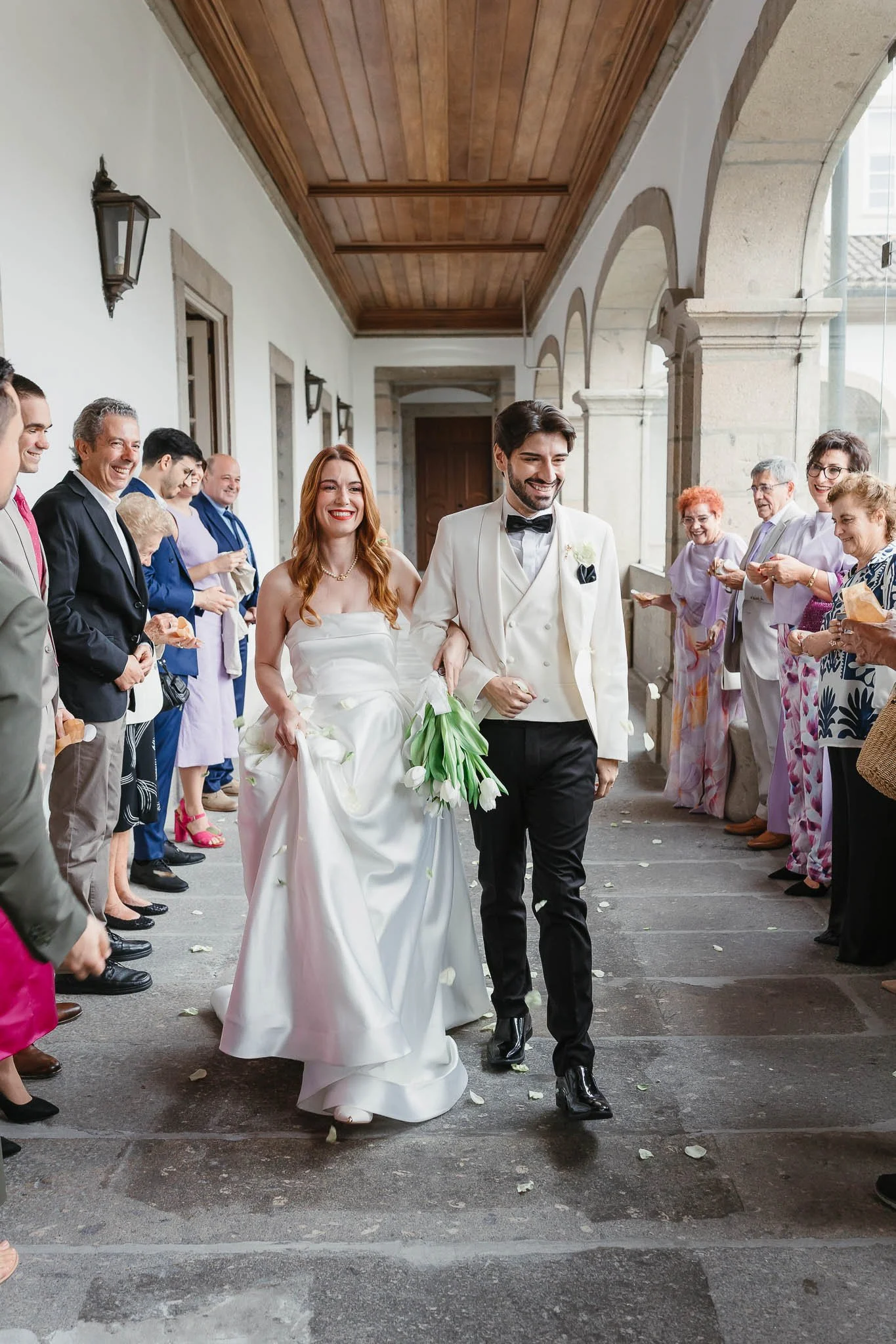 Portuguese Wedding Photographer:   Newlywed couple walking through wedding guests, holding flowers, surrounded by smiling guests, in an outdoor covered walkway.