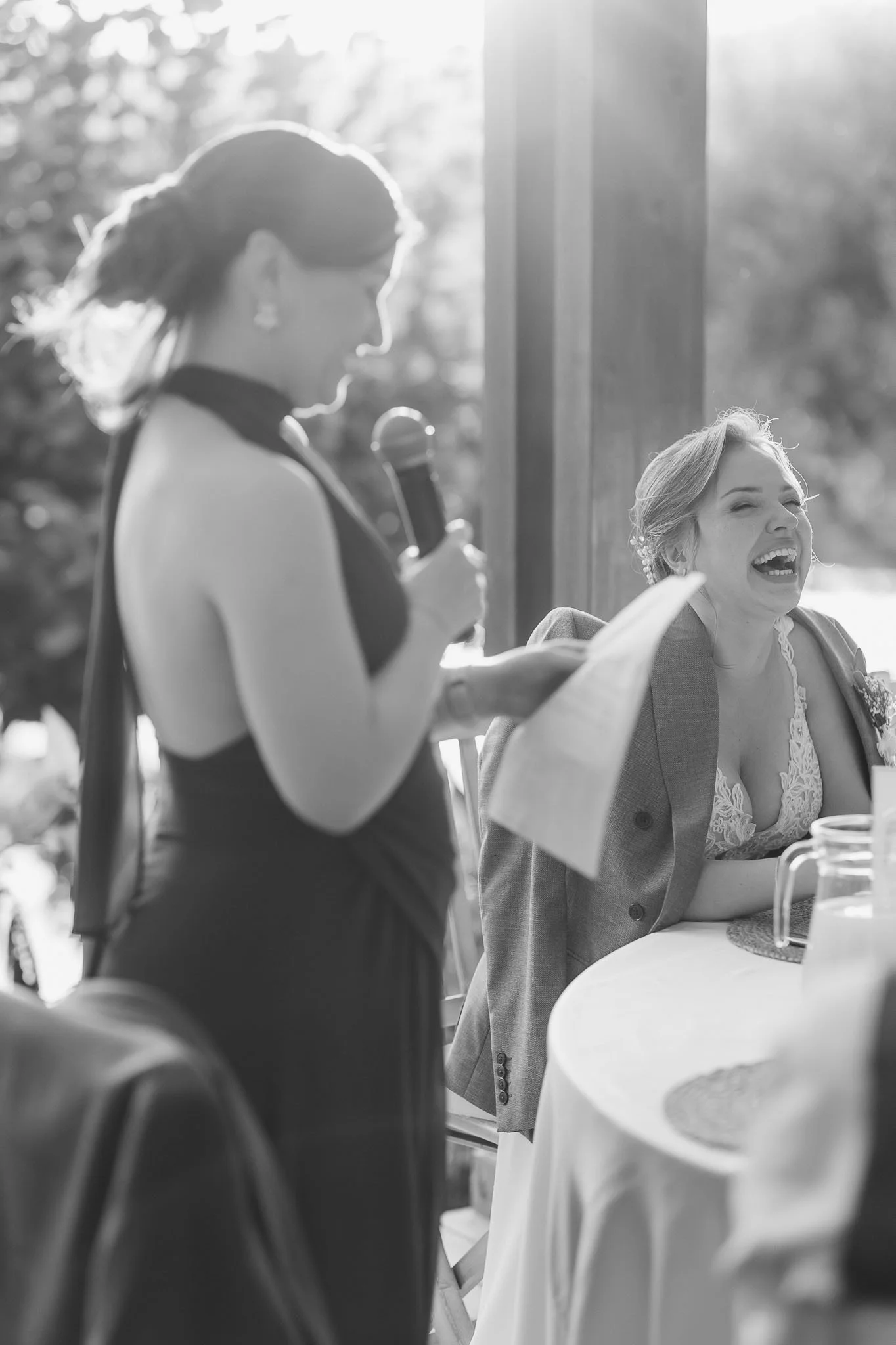 Emotional wedding moment in Portugal: A woman giving a speech while holding a microphone, and another woman laughing and smiling at a table during an outdoor event.