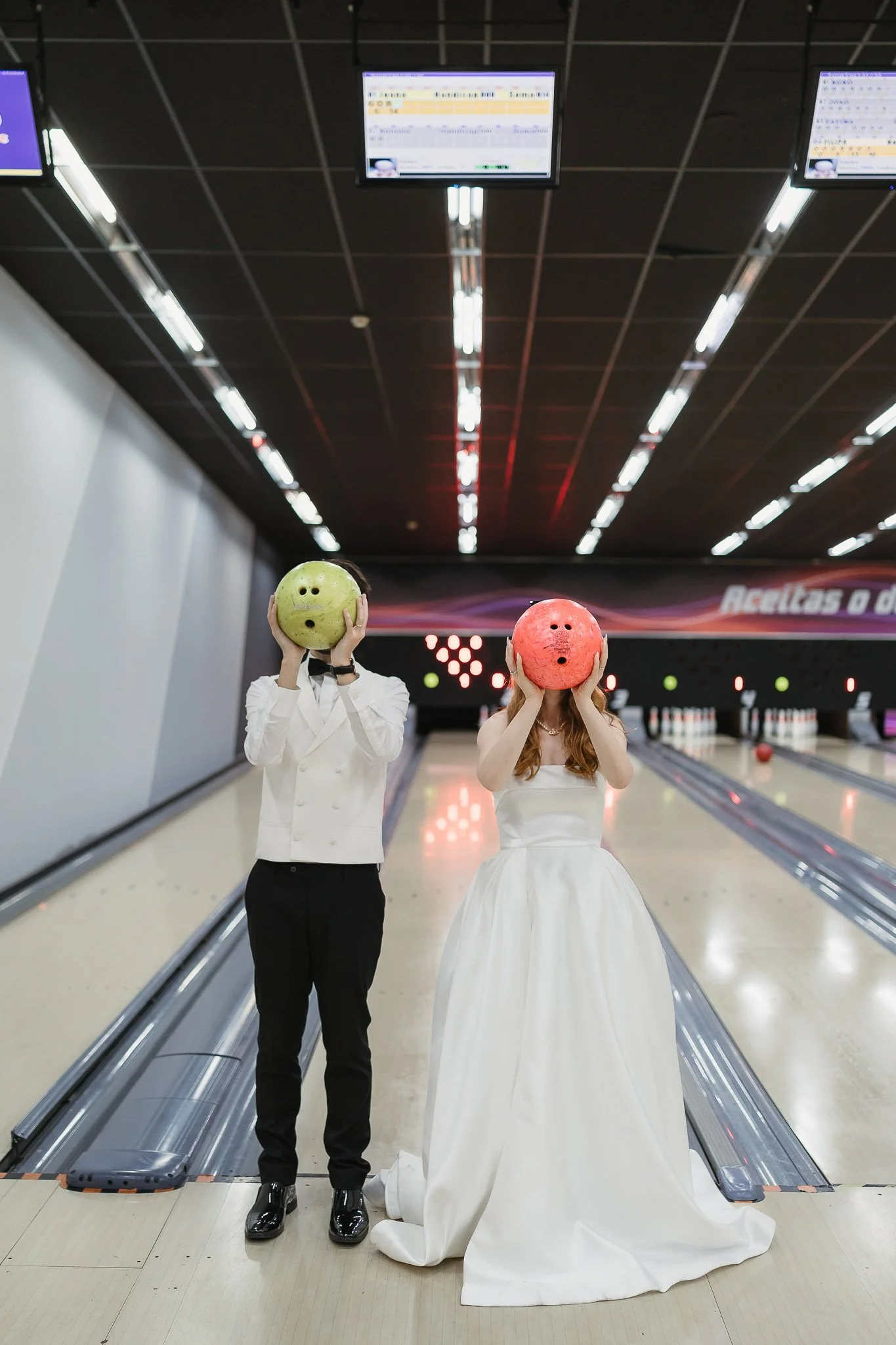 Two people in formal attire, one in a wedding dress and the other in a white jacket and black pants, standing at a bowling alley, holding bowling balls in front of their faces.