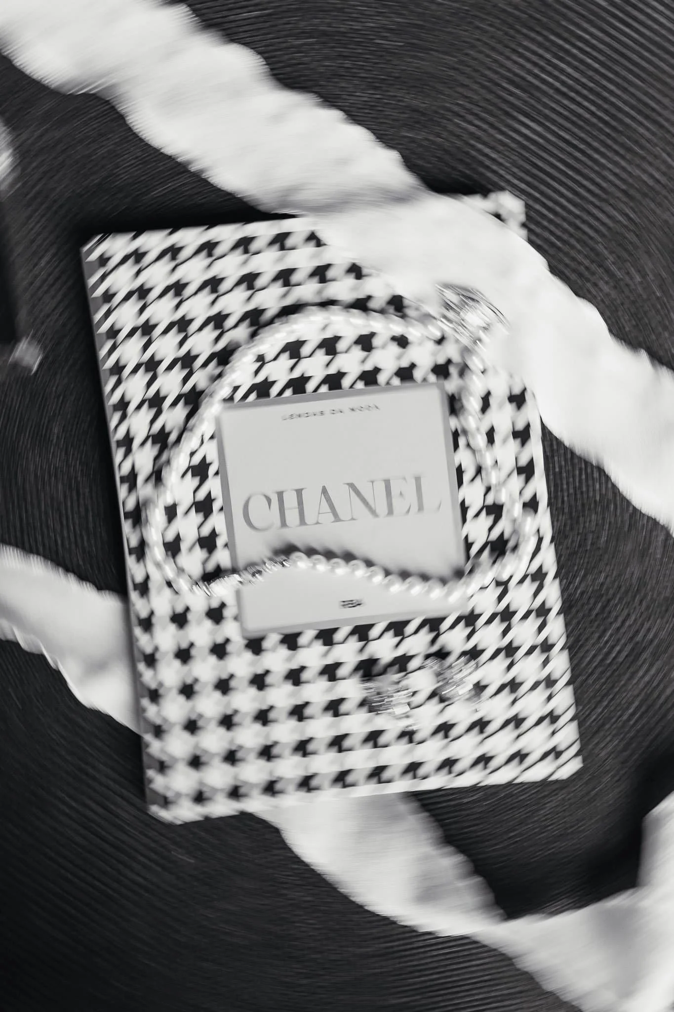 A Chanel jewelry box with pearl necklace on top, placed on a black and white houndstooth patterned bag, with white and black striped ribbon around the items.