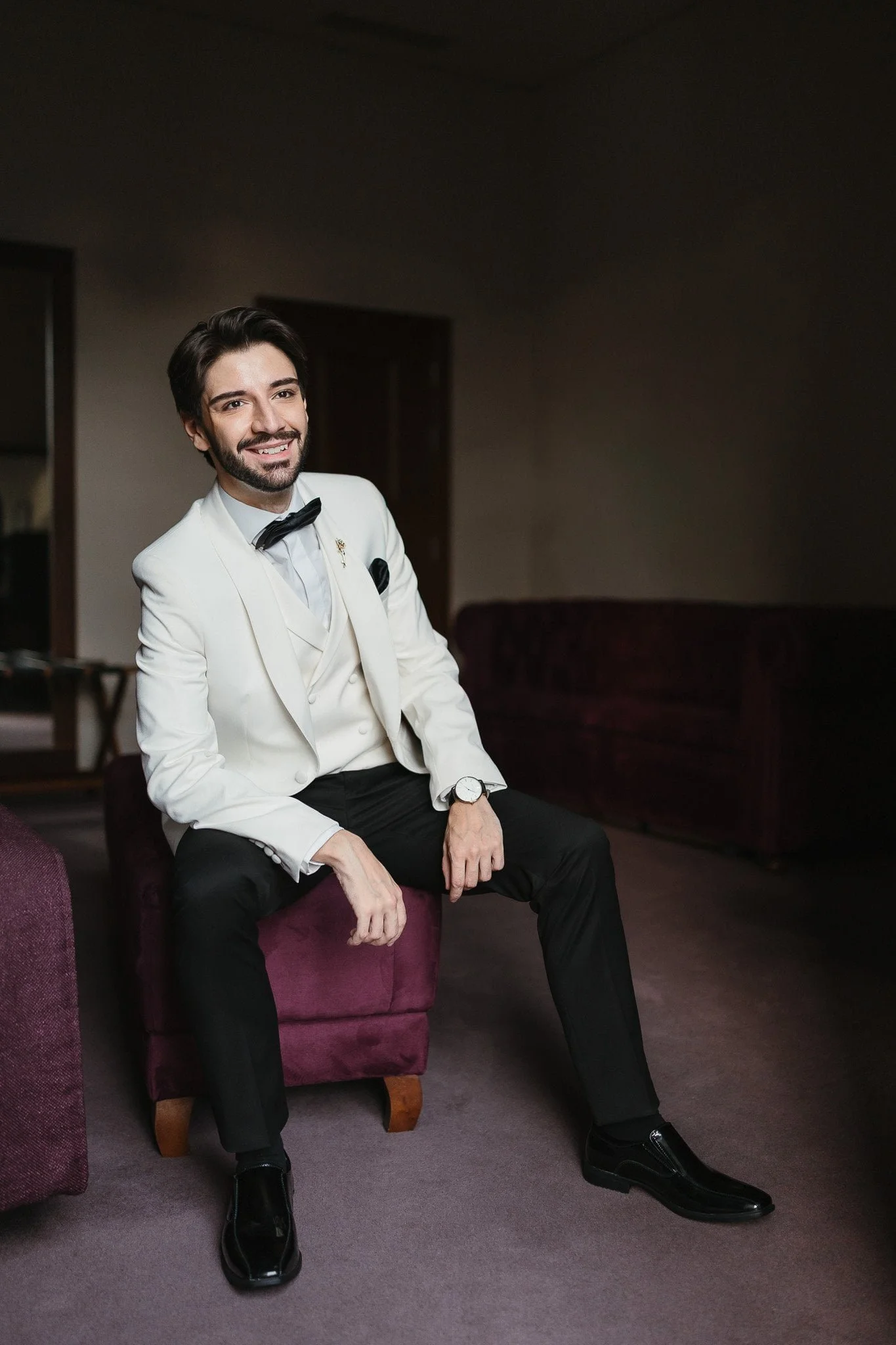 A groom dressed in a white tuxedo with a black bow tie, black pants, and black shoes, sitting on a purple upholstered ottoman in a dimly lit room, smiling at the camera. Portuguese Wedding Photographer