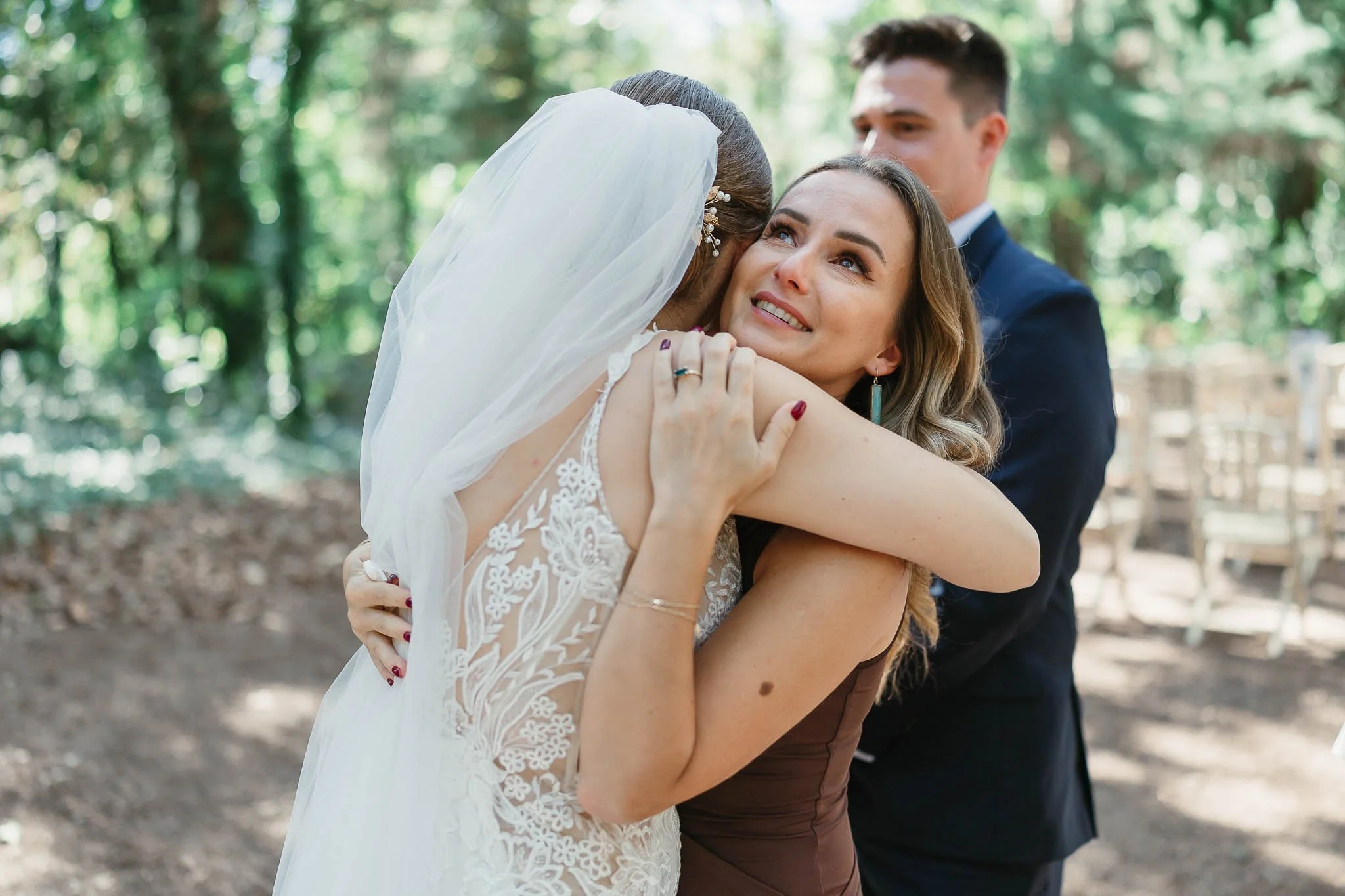A bride in a veil hugs a smiling woman outdoors, captured by a Portugal wedding photographer in a wooded setting.