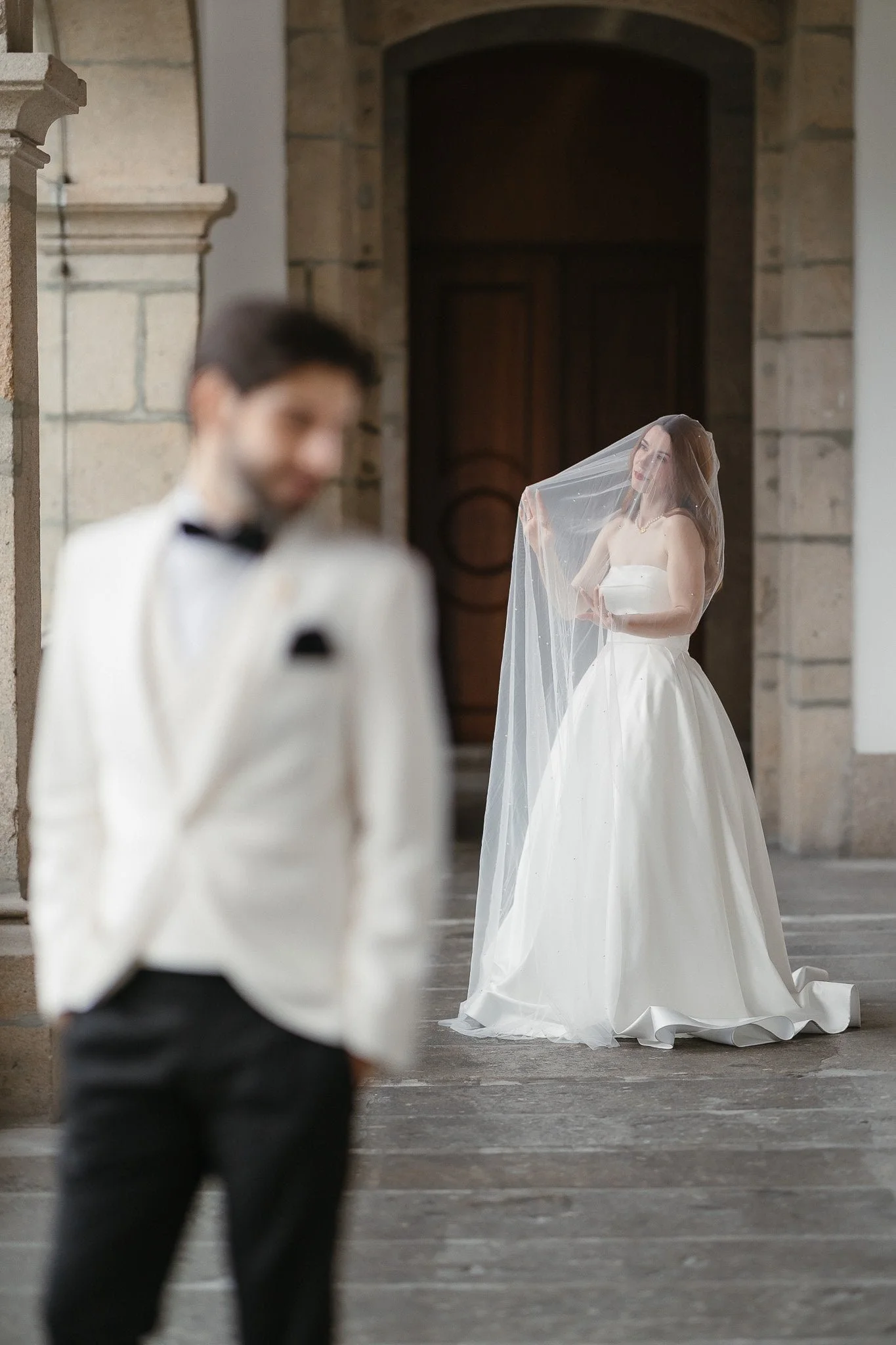 A bride in a white gown with a veil looks at a groom in a tuxedo, with a blurred focus in the foreground, inside a stone building.