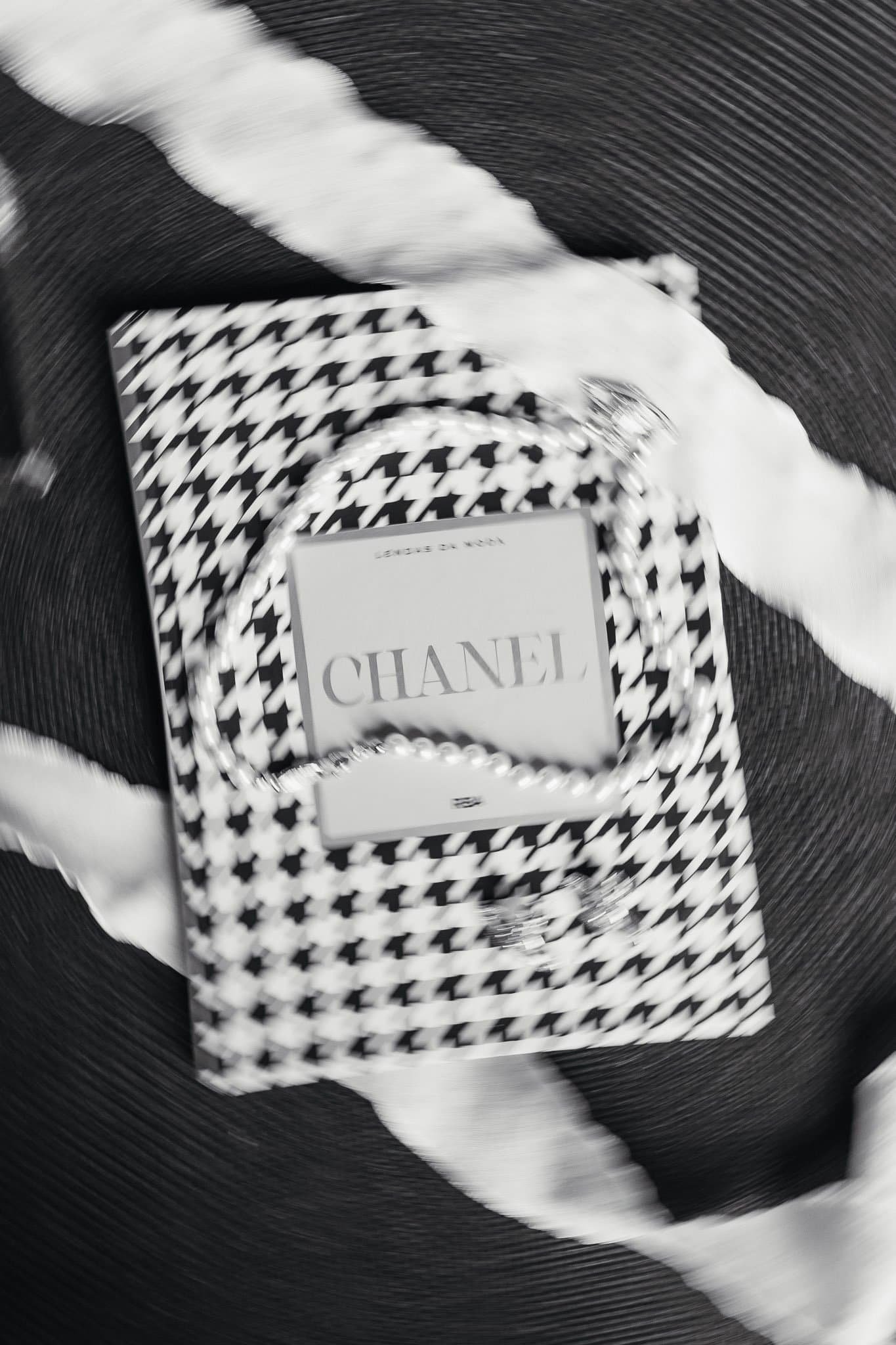 A Chanel jewelry box with pearl necklace on top, placed on a black and white houndstooth patterned bag, with white and black striped ribbon around the items.