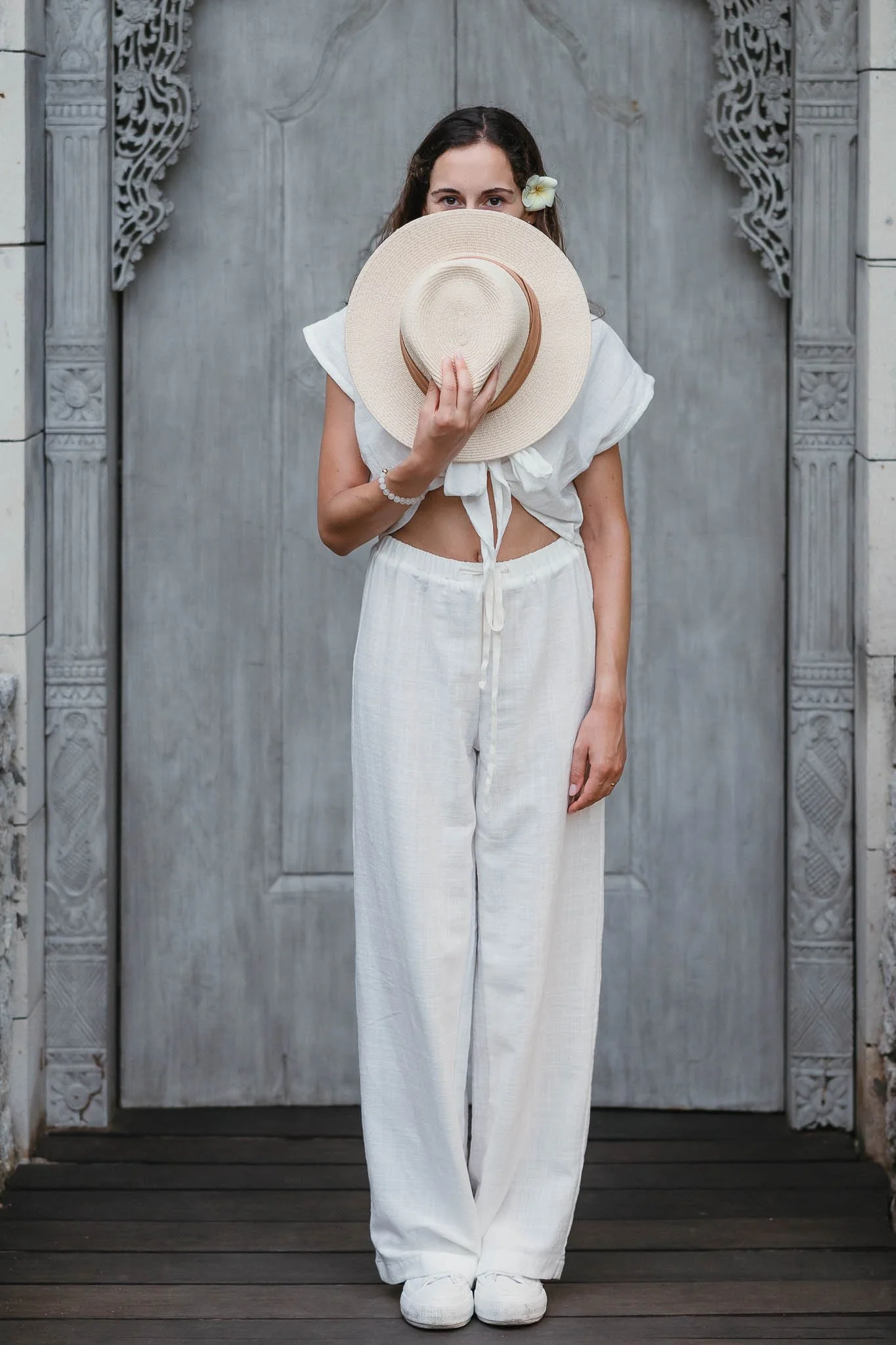 A woman standing in front of an ornate gray wooden door, holding a large straw hat to her face with one hand, while the other hand rests by her side. She has a flower in her hair and is dressed in a white, tied crop top and loose white pants.
