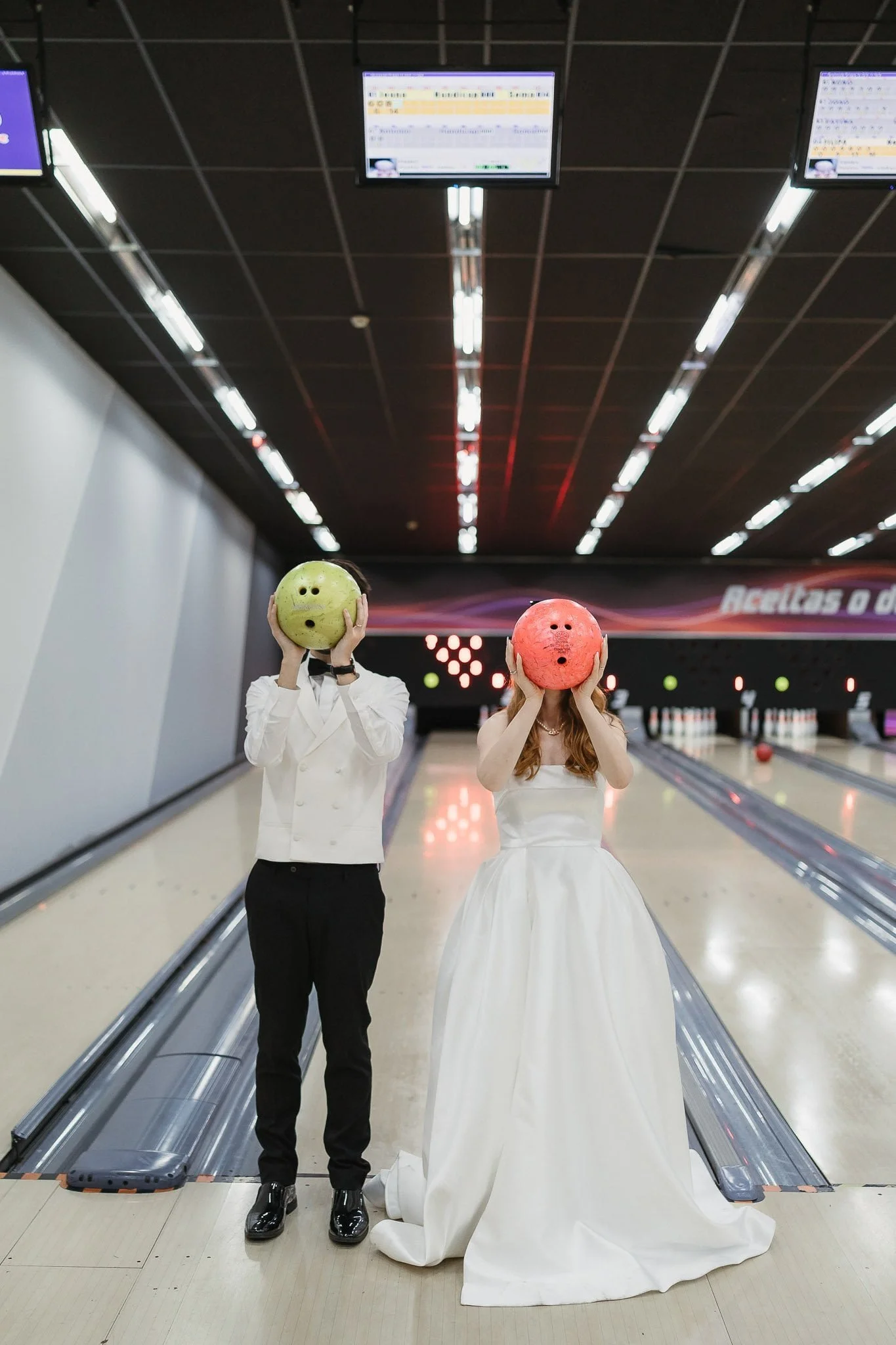 Two people in formal attire, one in a wedding dress and the other in a white jacket and black pants, standing at a bowling alley, holding bowling balls in front of their faces.
