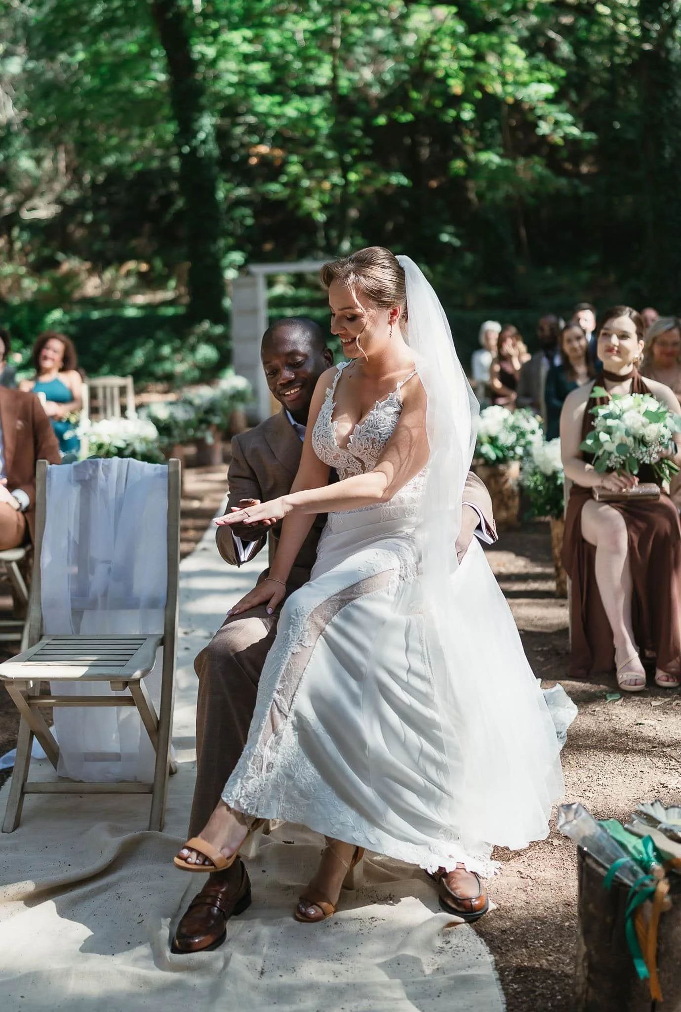 A bride in a white lace wedding dress and veil sitting on a man's lap during a wedding ceremony outdoors. Guests seated behind them in a forest setting, some holding bouquets, watching and smiling.