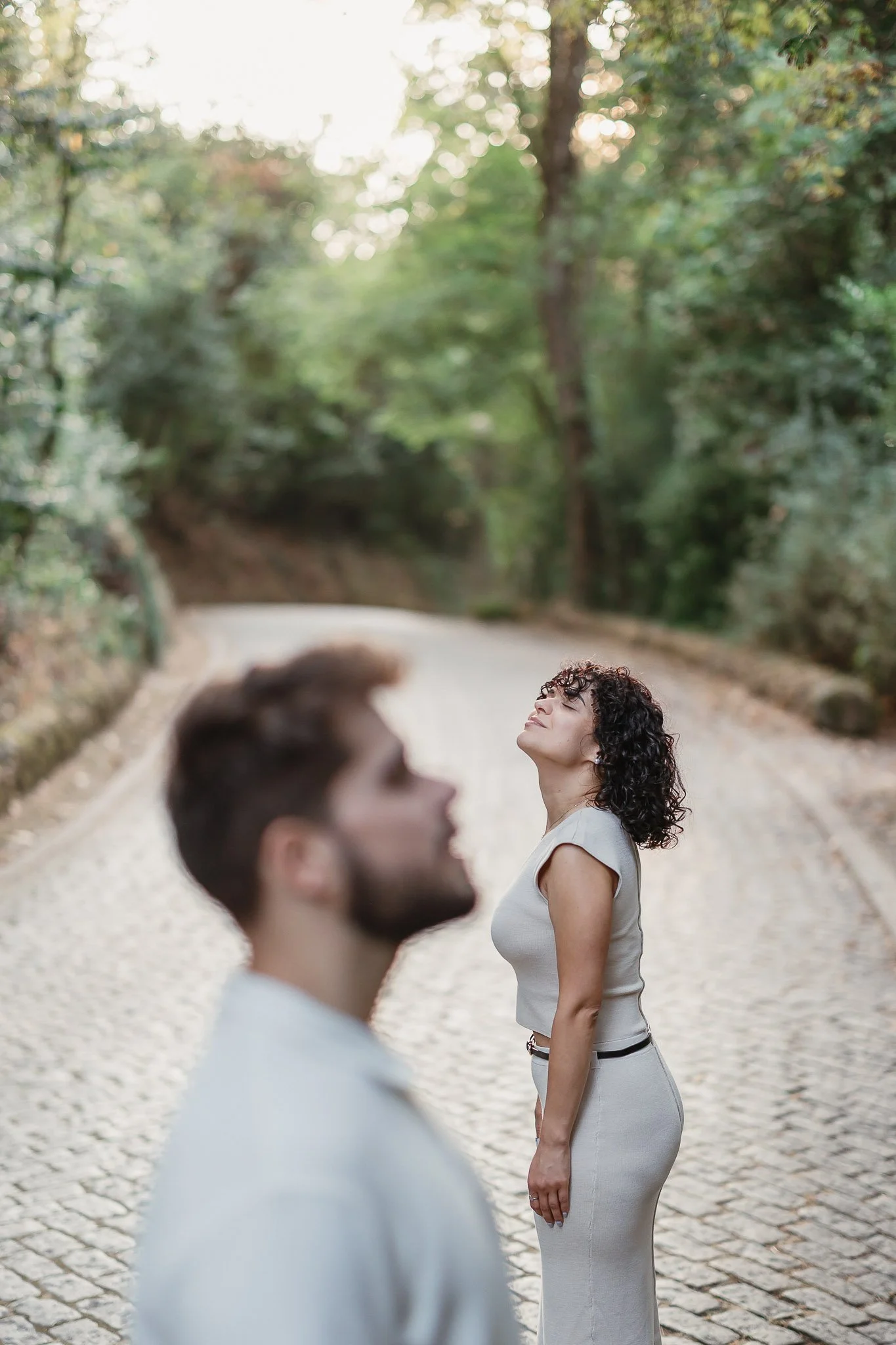 A woman with curly hair standing on a cobblestone path surrounded by trees, looking up with a serene expression, while a man with short hair and beard stands in the foreground with his face blurred.
