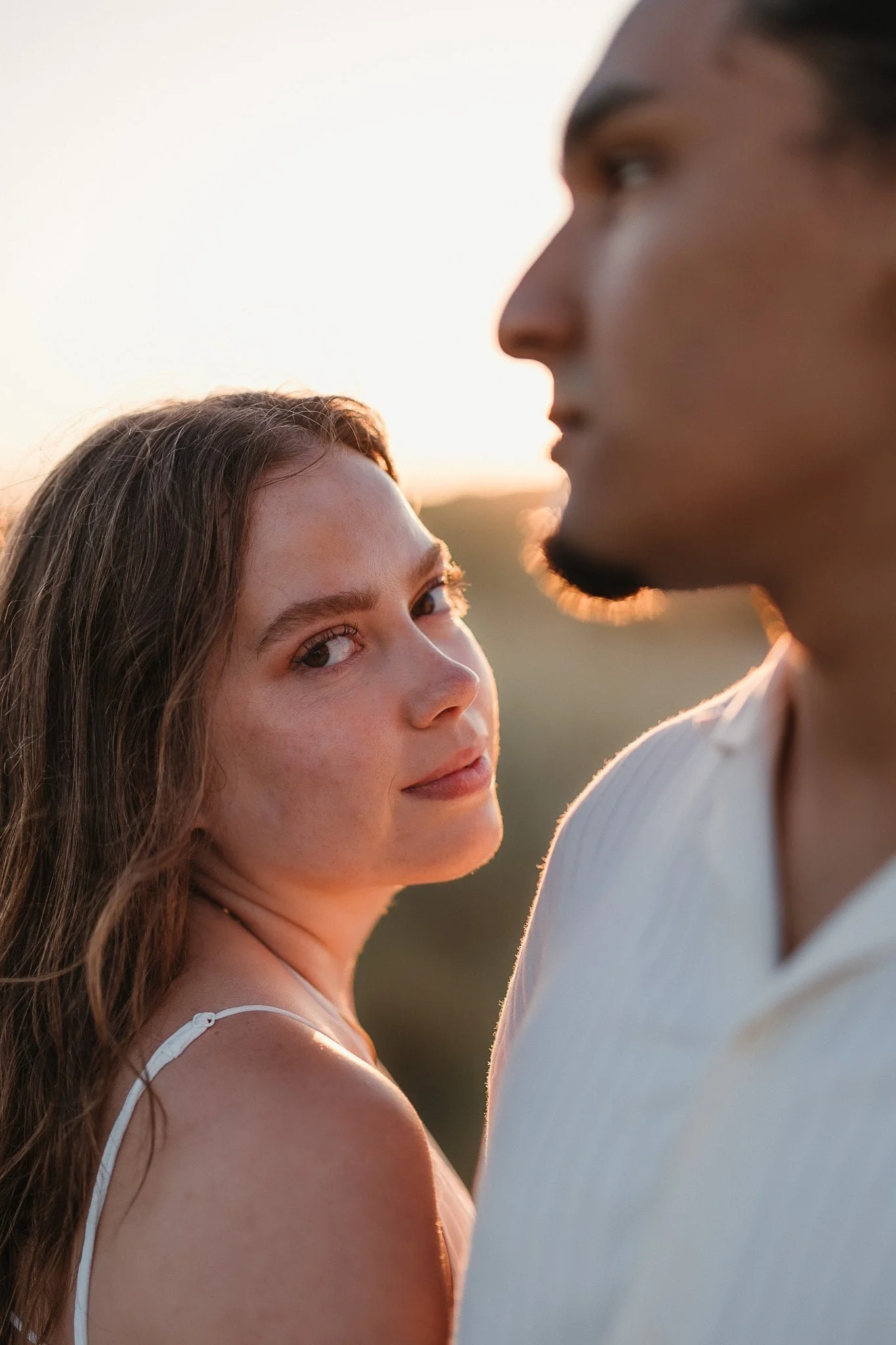 A woman gazes at the camera over a man's shoulder at sunset—light attire, outdoors. Captured by a Portugal wedding photographer. Engagement Session