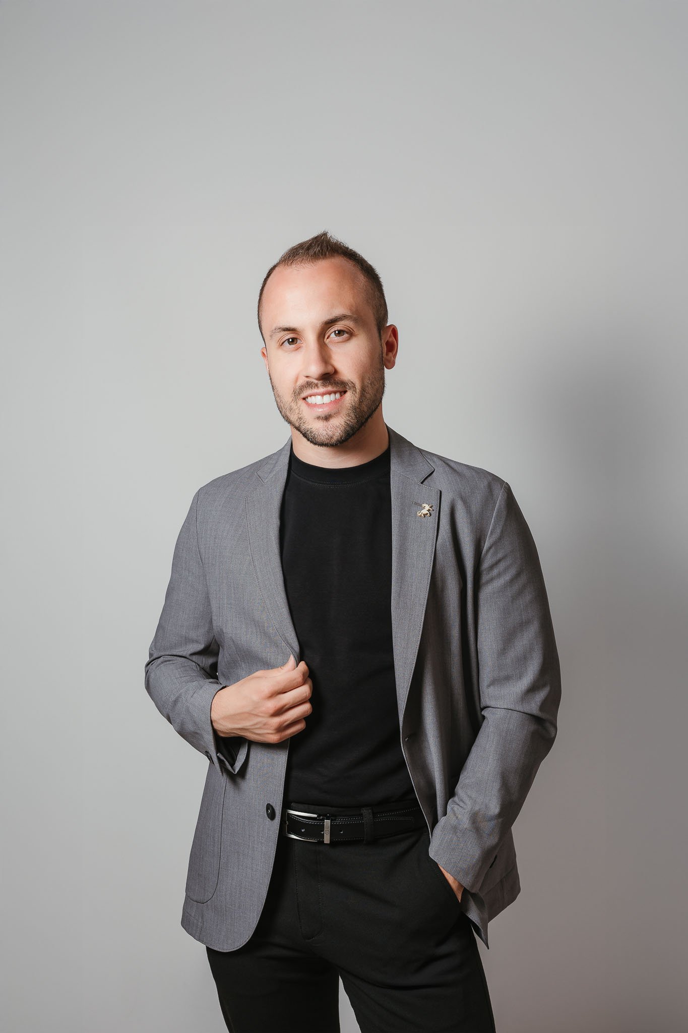 A young man with short hair and a beard wearing a gray blazer over a black shirt, smiling and standing against a plain light gray background.