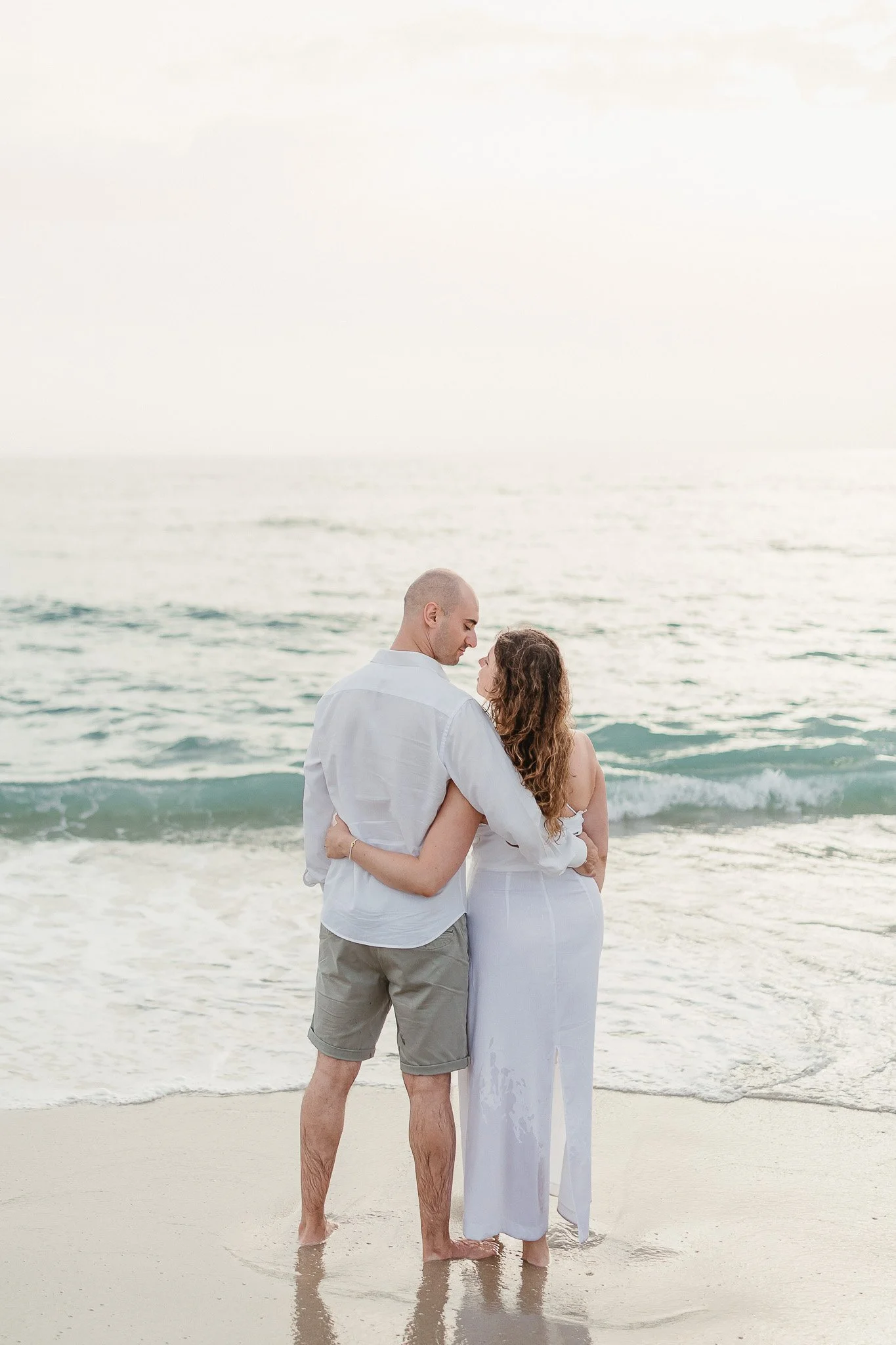 A couple embraces on a sandy beach, gazing at the ocean under a cloudy sky—captured by a Portugal wedding photographer.
