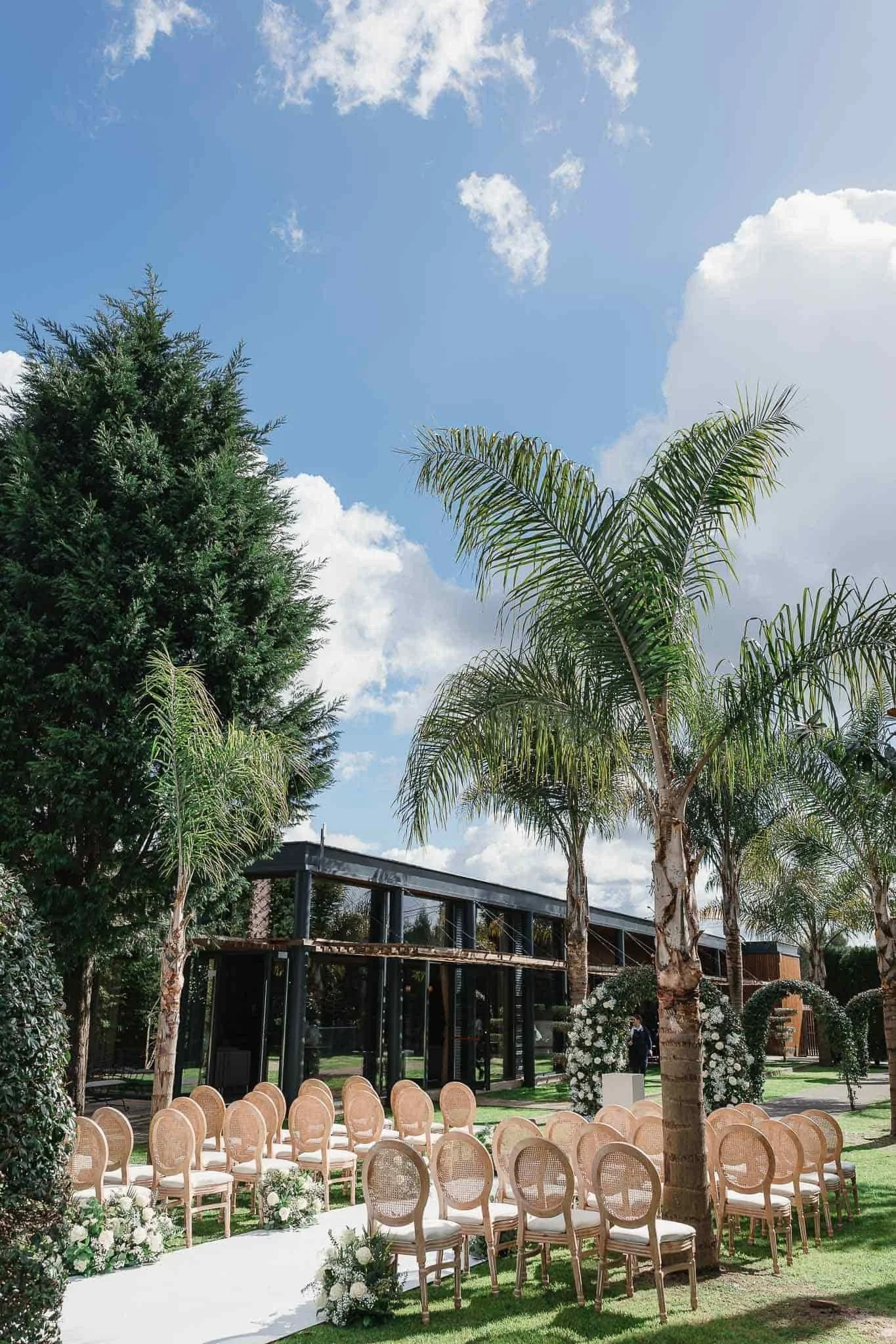 Outdoor wedding setup with rows of chairs, floral arrangements, palm trees, and a modern building in the background under a partly cloudy sky.