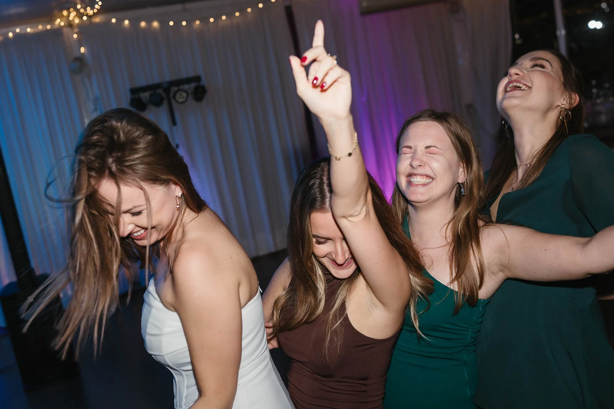 Four young women in dresses laugh and dance at a party—captured by a Portugal Wedding Photographer under glowing string lights.