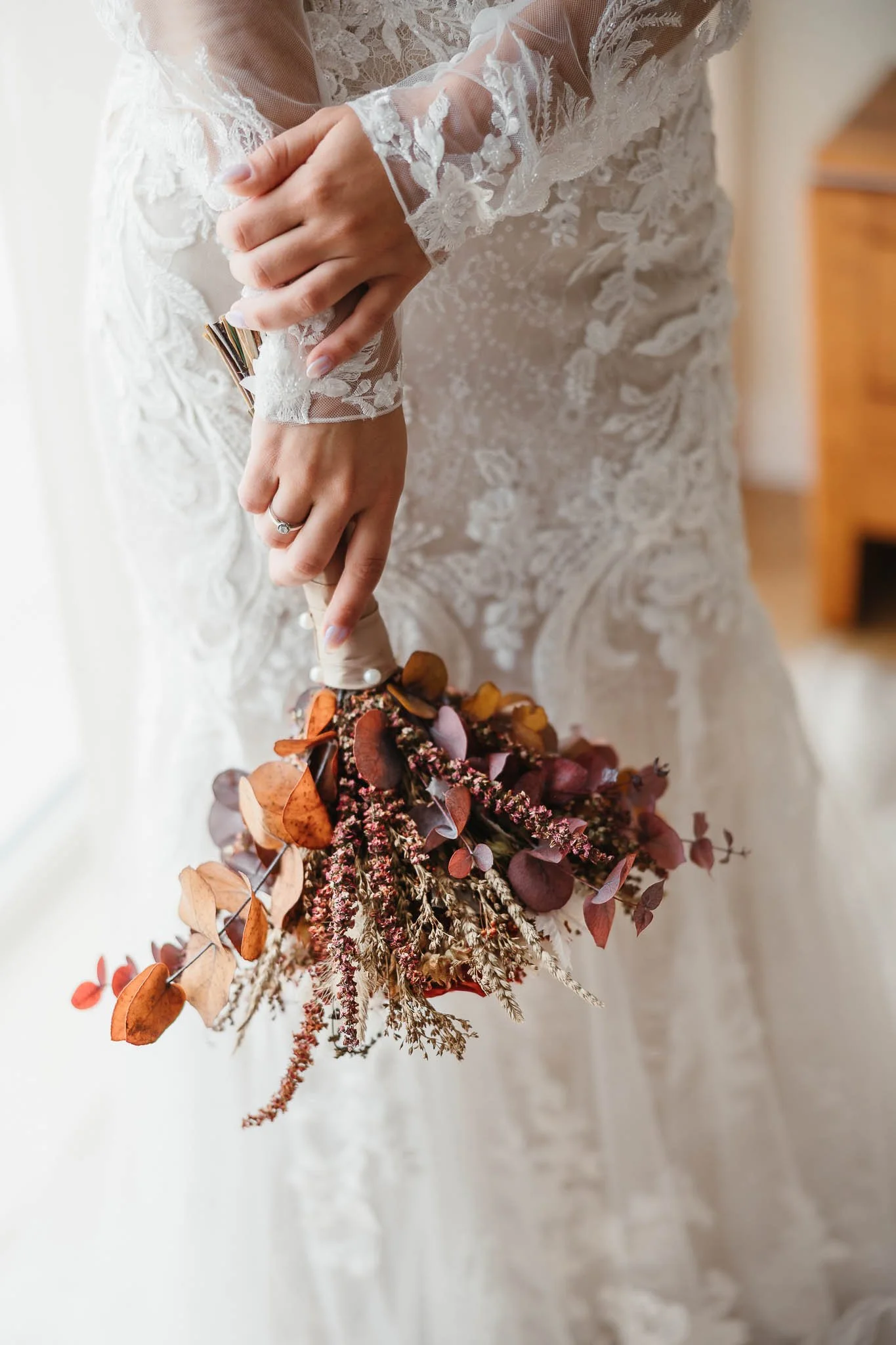 Close-up of a bride holding a bouquet of dried flowers, including leaves and twigs, with her hands in a lace wedding dress.