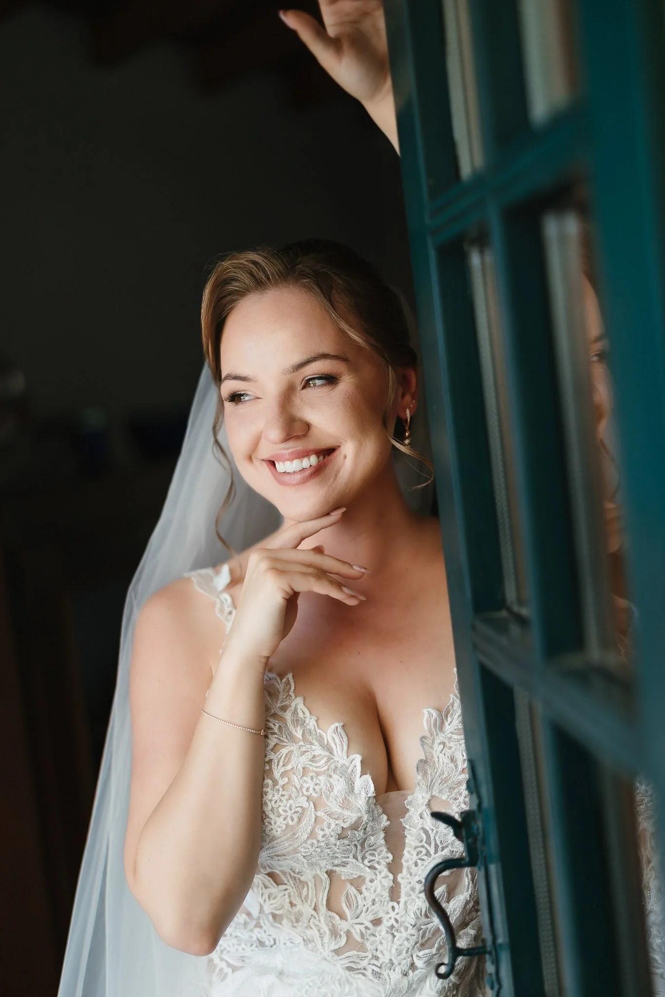 A smiling bride in a white lace wedding dress looking out a window, with her hand resting on her chin.