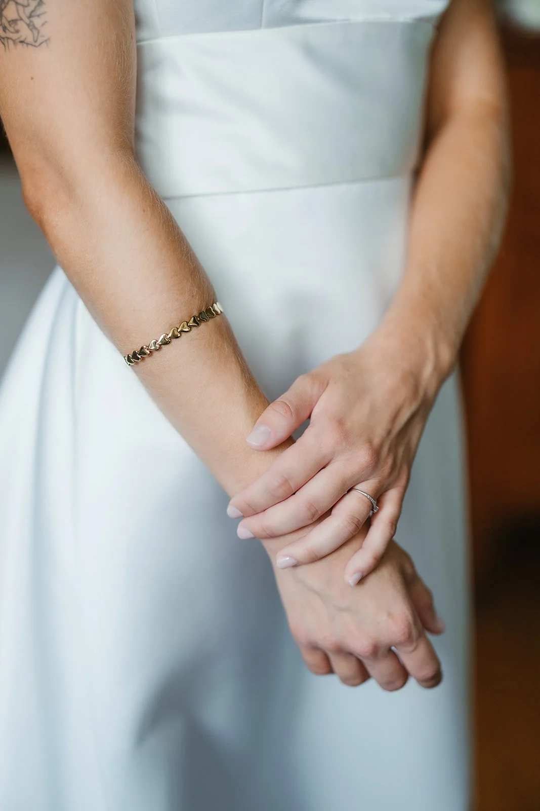 A bride in a white dress stands with hands gently crossed, showcasing a gold bracelet and ring—Portugal Wedding Photography.