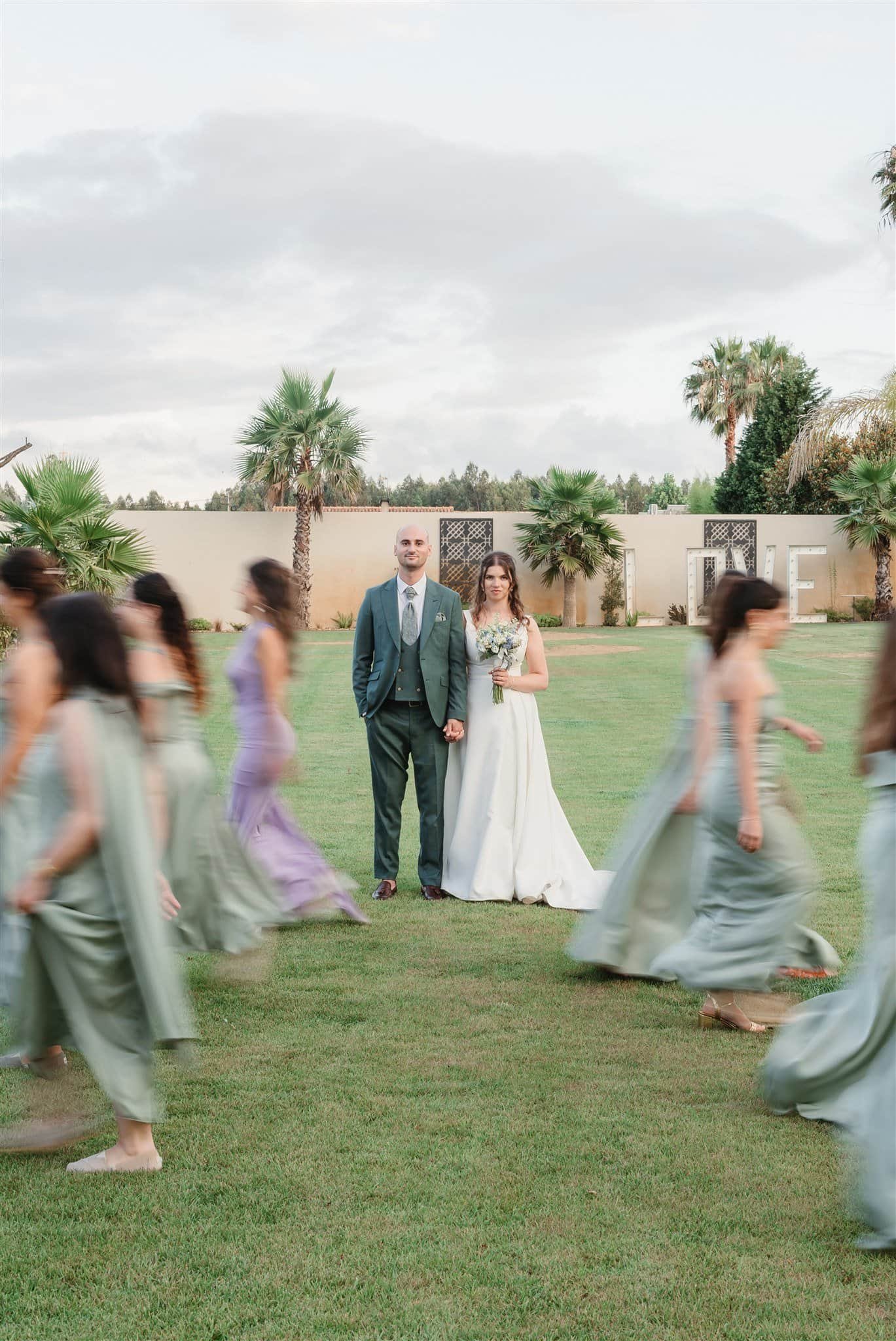 A bride and groom standing together on a grassy field with palm trees and a wall with large decorative letters spelling 'LOVE' in the background, surrounded by women in pastel-colored dresses walking in a circle.