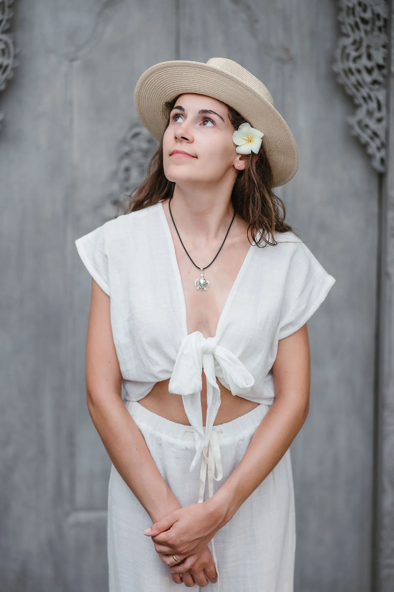 A young woman wearing a straw hat with a white flower, a white tied-up crop top, and matching white pants, standing against a gray wooden background, looking upwards.