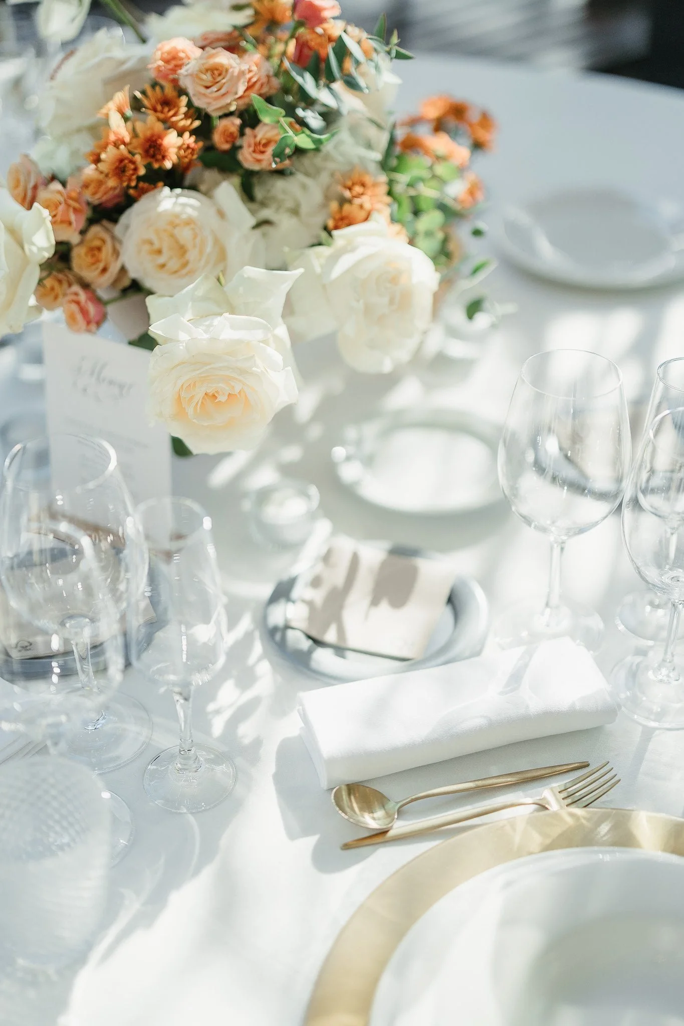 Elegant table setting with a white tablecloth, glassware, a gold-rimmed plate, gold utensils, a white napkin, and a floral centerpiece of white and pink roses and greenery - Wedding Photography Portugal