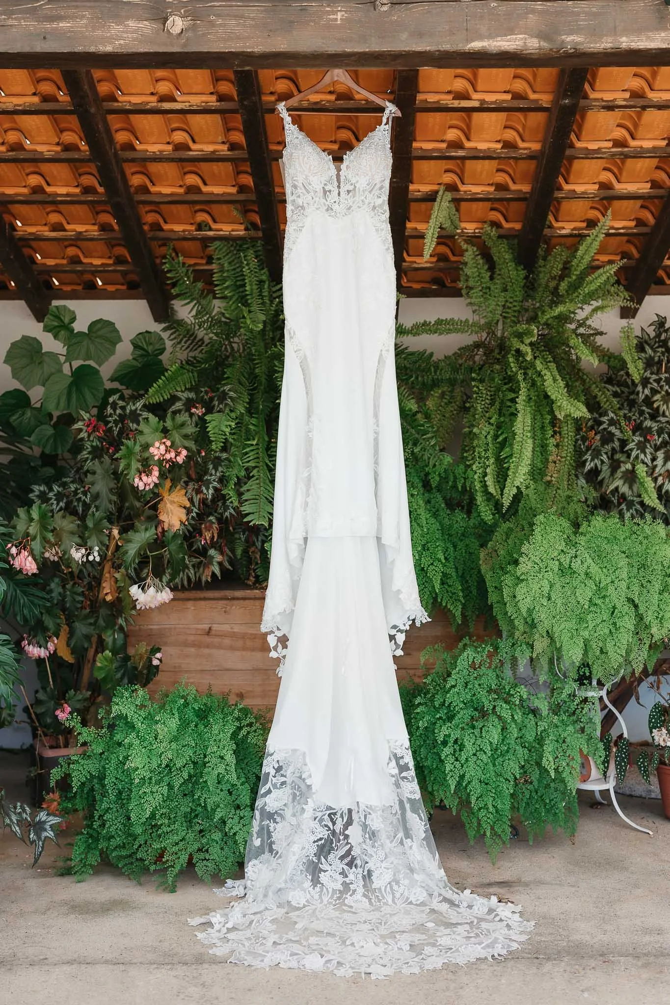 White lace wedding dress hanging from a hanger against a background of green plants and a wooden ceiling with orange tiles.