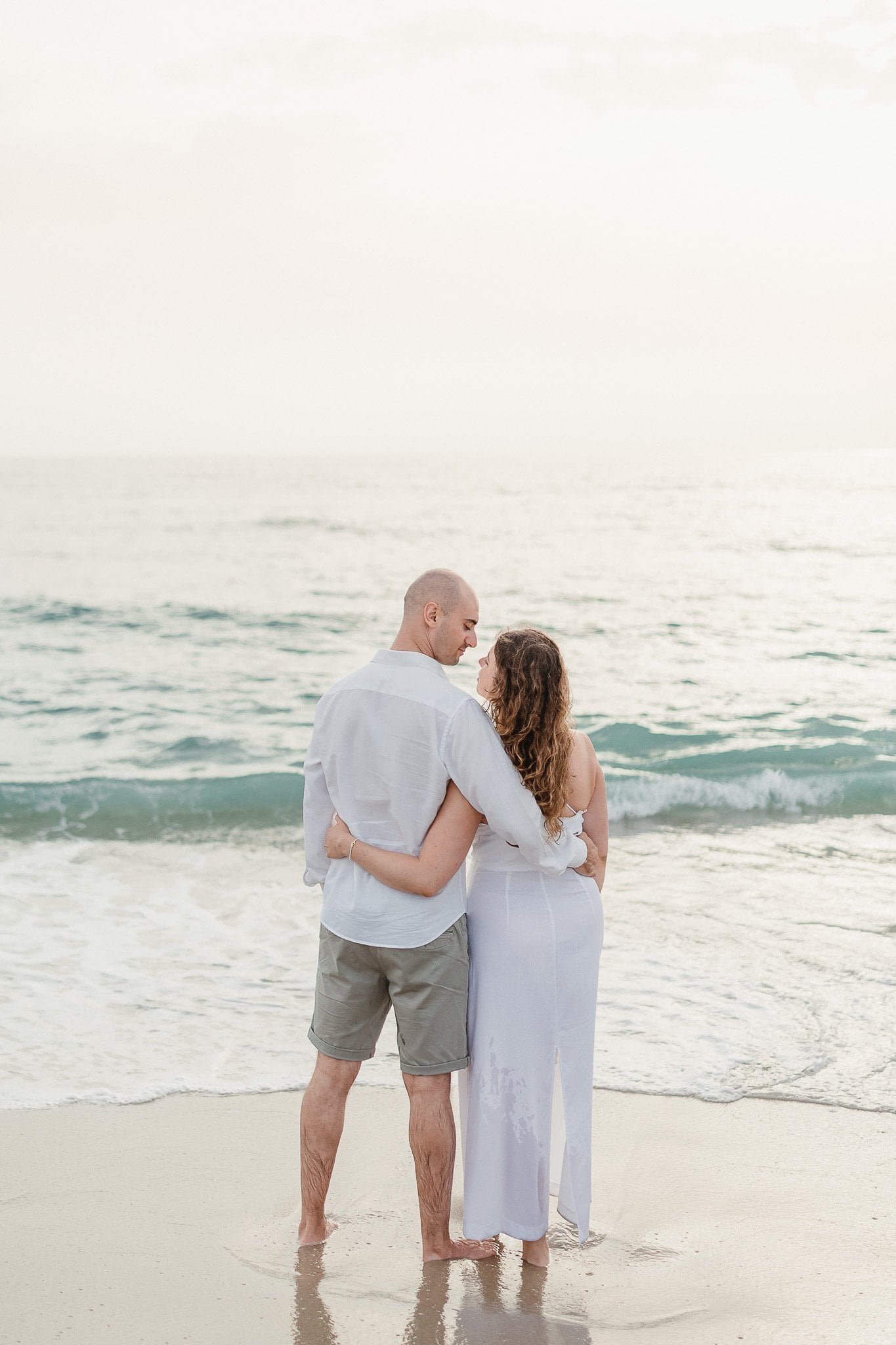 A couple stands arm in arm on the beach, facing the ocean, captured by a Portugal wedding photographer in soft, golden light.