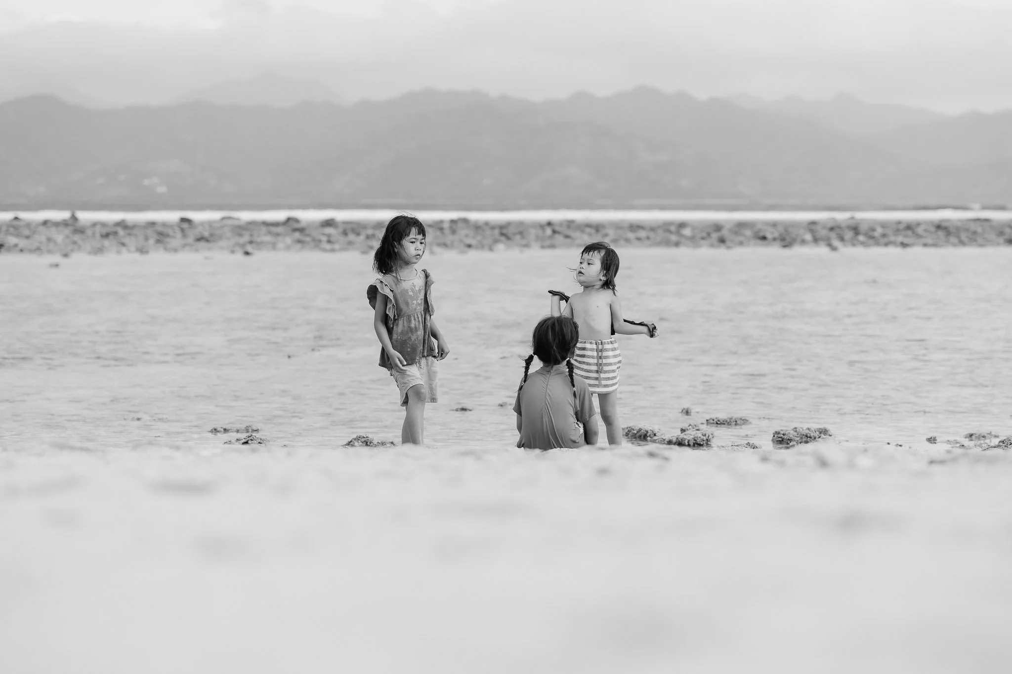 Three children playing on the beach, two girls standing and one girl sitting, with mountains and water in the background, in black and white.