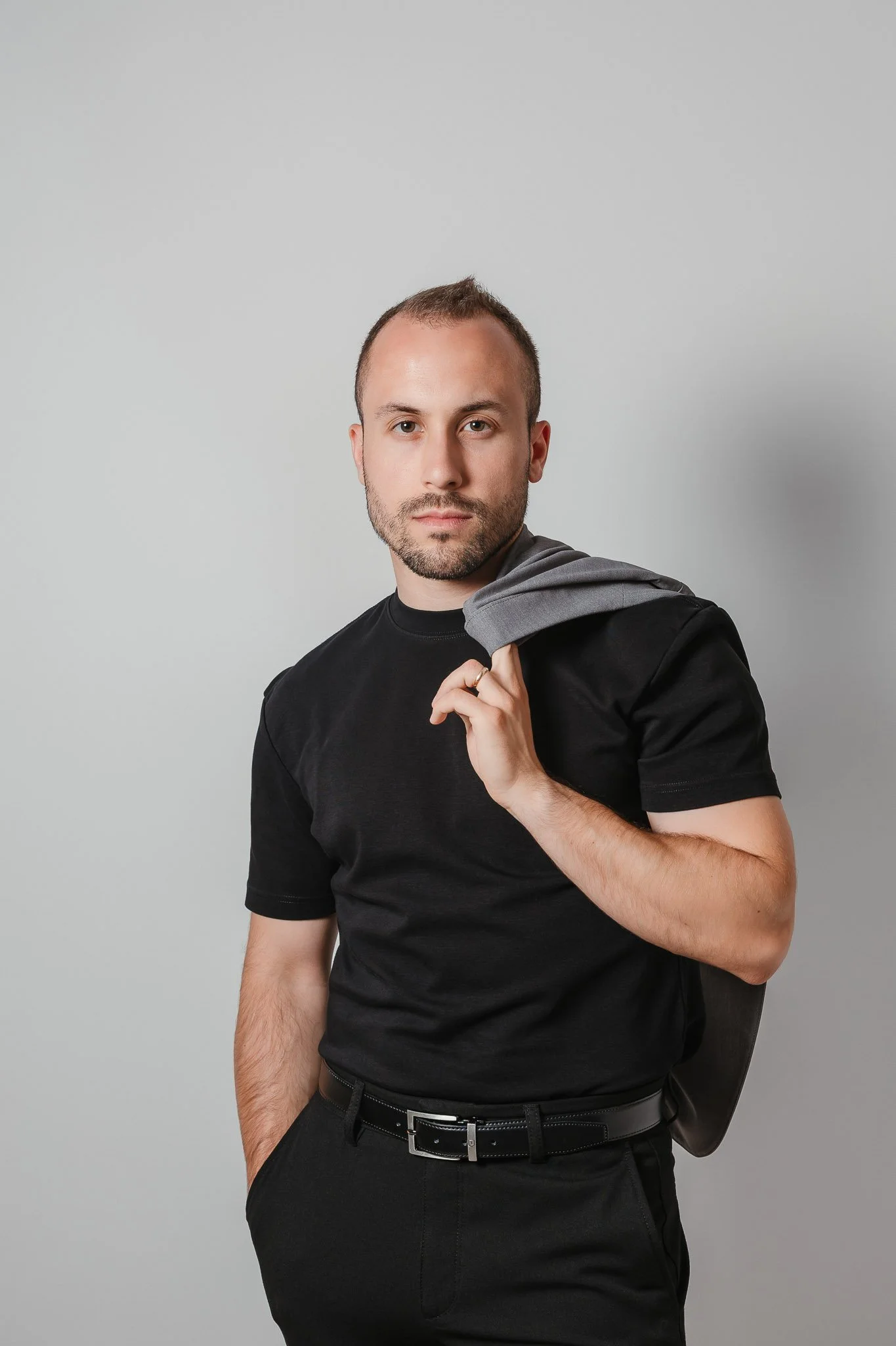 Fotógrafo de Casamentos em Portugal: Retrato de um jovem com cabelo curto e barba, vestindo t-shirt preta, segurando um casaco cinza sobre o ombro, posando contra um fundo cinza simples.