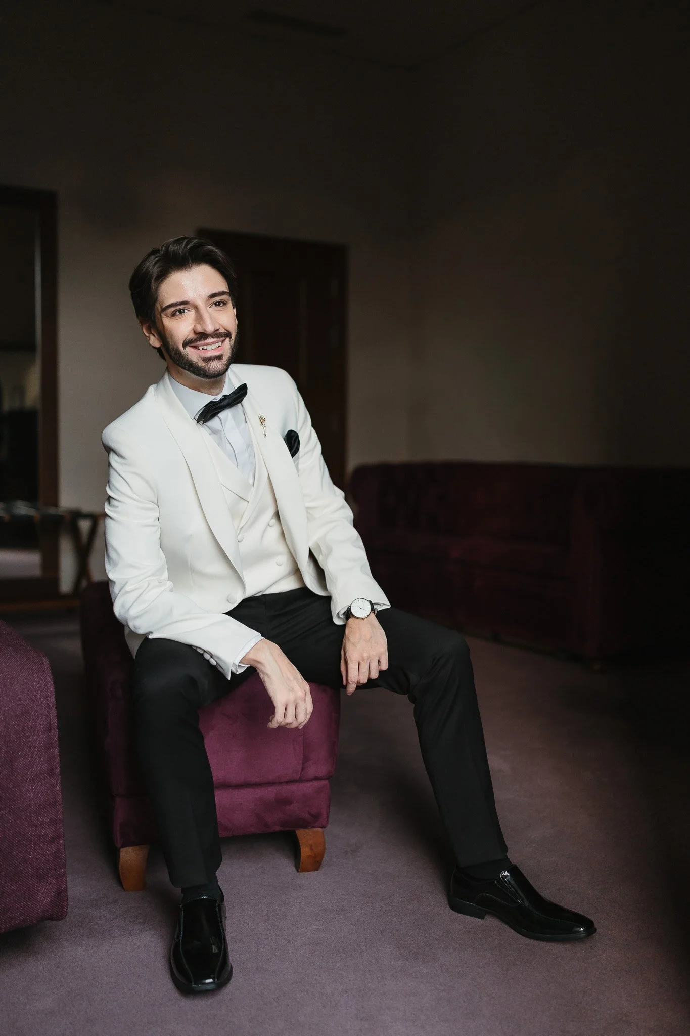 A groom dressed in a white tuxedo with a black bow tie, black pants, and black shoes, sitting on a purple upholstered ottoman in a dimly lit room, smiling at the camera. Portuguese Wedding Photographer
