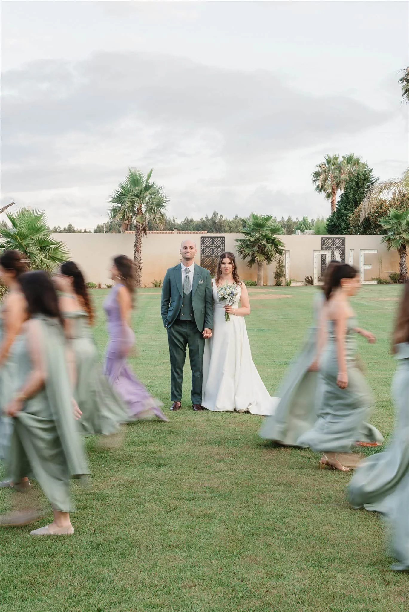 A bride and groom standing together on a grassy field with palm trees and a wall with large decorative letters spelling 'LOVE' in the background, surrounded by women in pastel-colored dresses walking in a circle. -