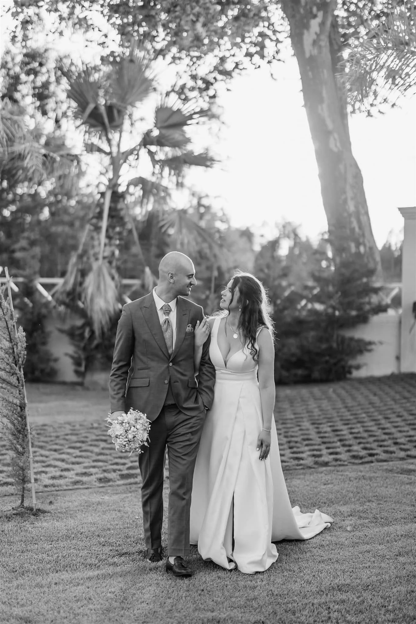 Bride and groom walk hand in hand outdoors, smiling in a garden. Black and white photo by Portugal Wedding Photographer.