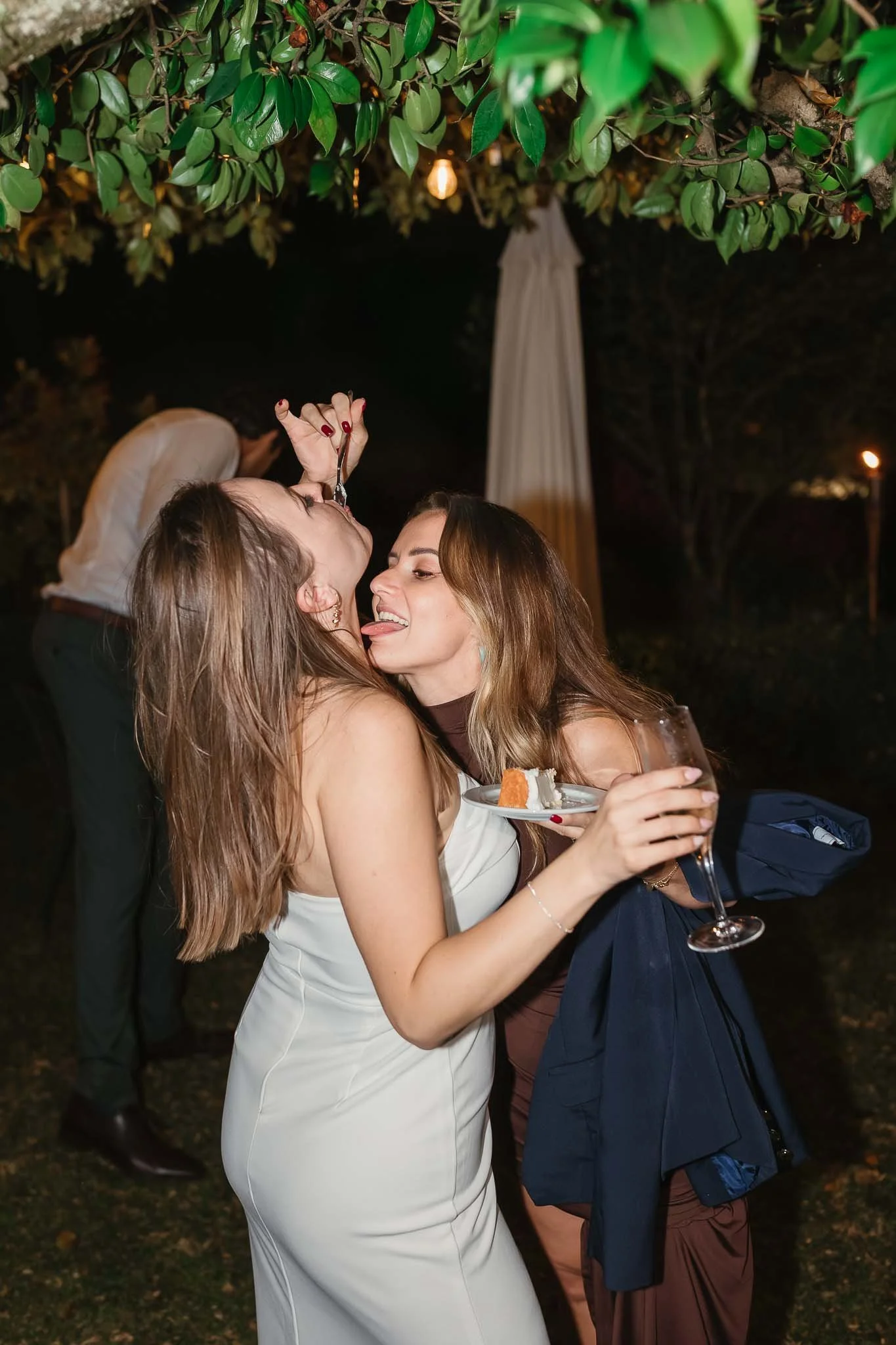 Two women in dresses laugh and enjoy dessert under greenery at an outdoor night party—captured by a Portugal Wedding Photographer.