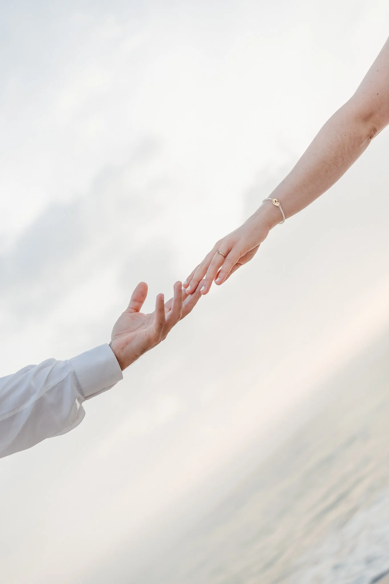 Engament Sesion in Aveiro: Close-up of two people reaching out towards each other, one with a white shirt and the other with a wrist bracelet and rings, with a cloudy sky and water in the background.