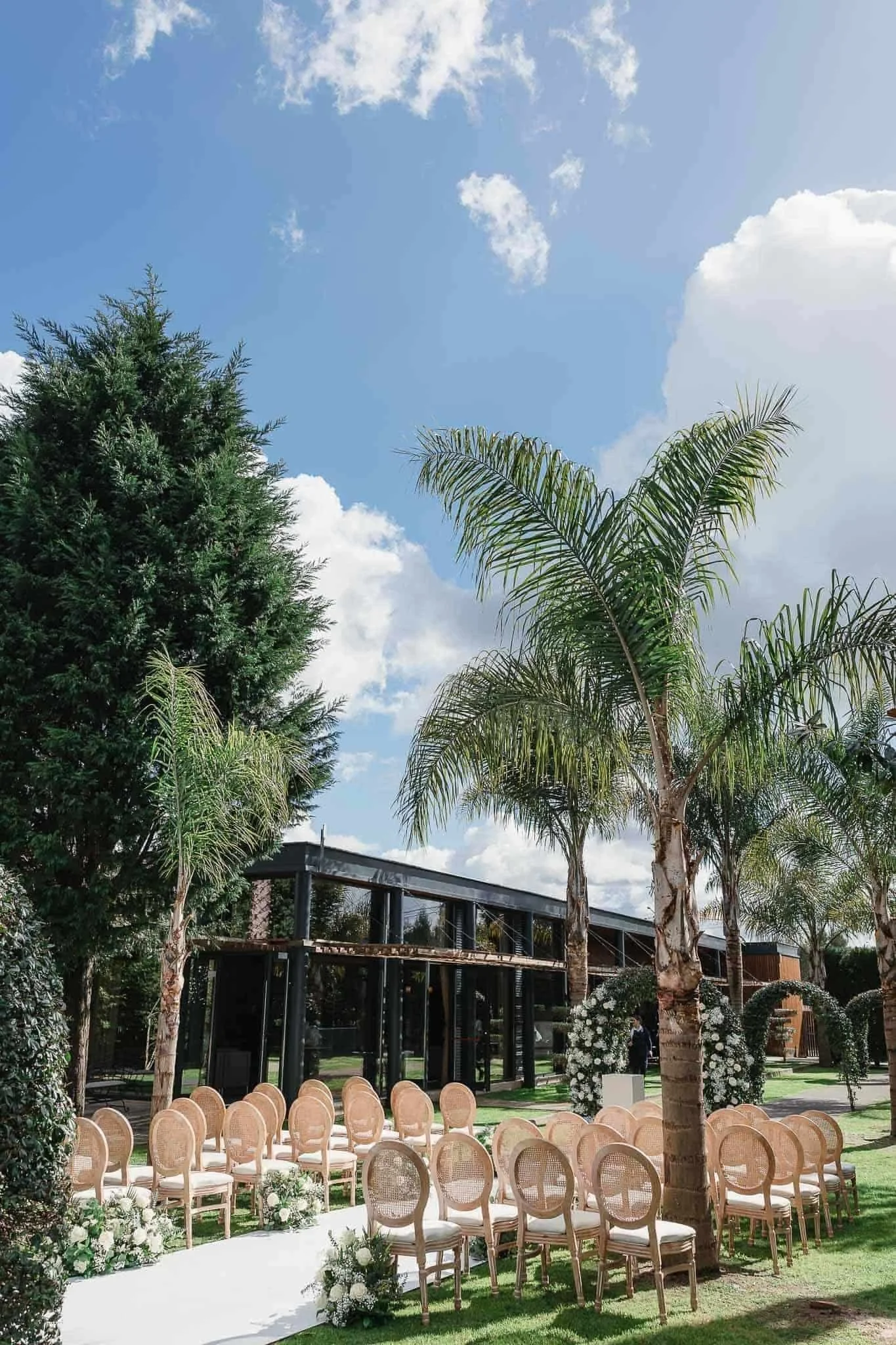 Destination Wedding Photographer Portugal - Outdoor wedding setup with rows of chairs, floral arrangements, palm trees, and a modern building in the background under a partly cloudy sky.